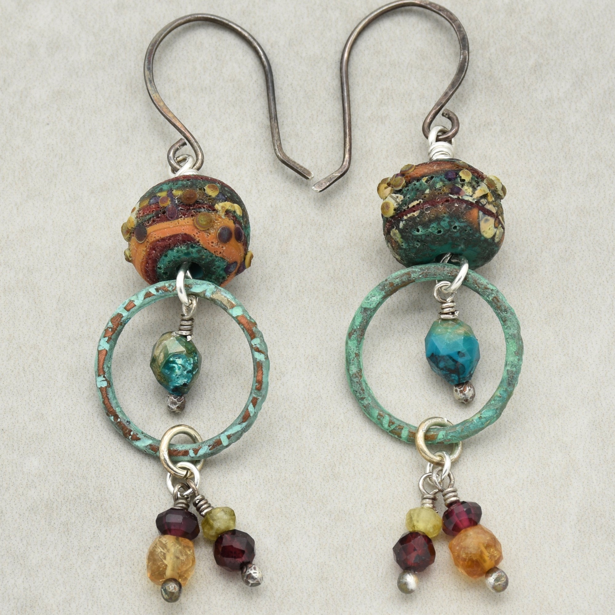 Unique autumn tone handmade pair of earrings with artisan lampwork glass, chrysocolla, green garnet, red garnet, purple garnet, citrine, oxidized silver ear wires, and artisan patinated turquoise rings on a light gray background