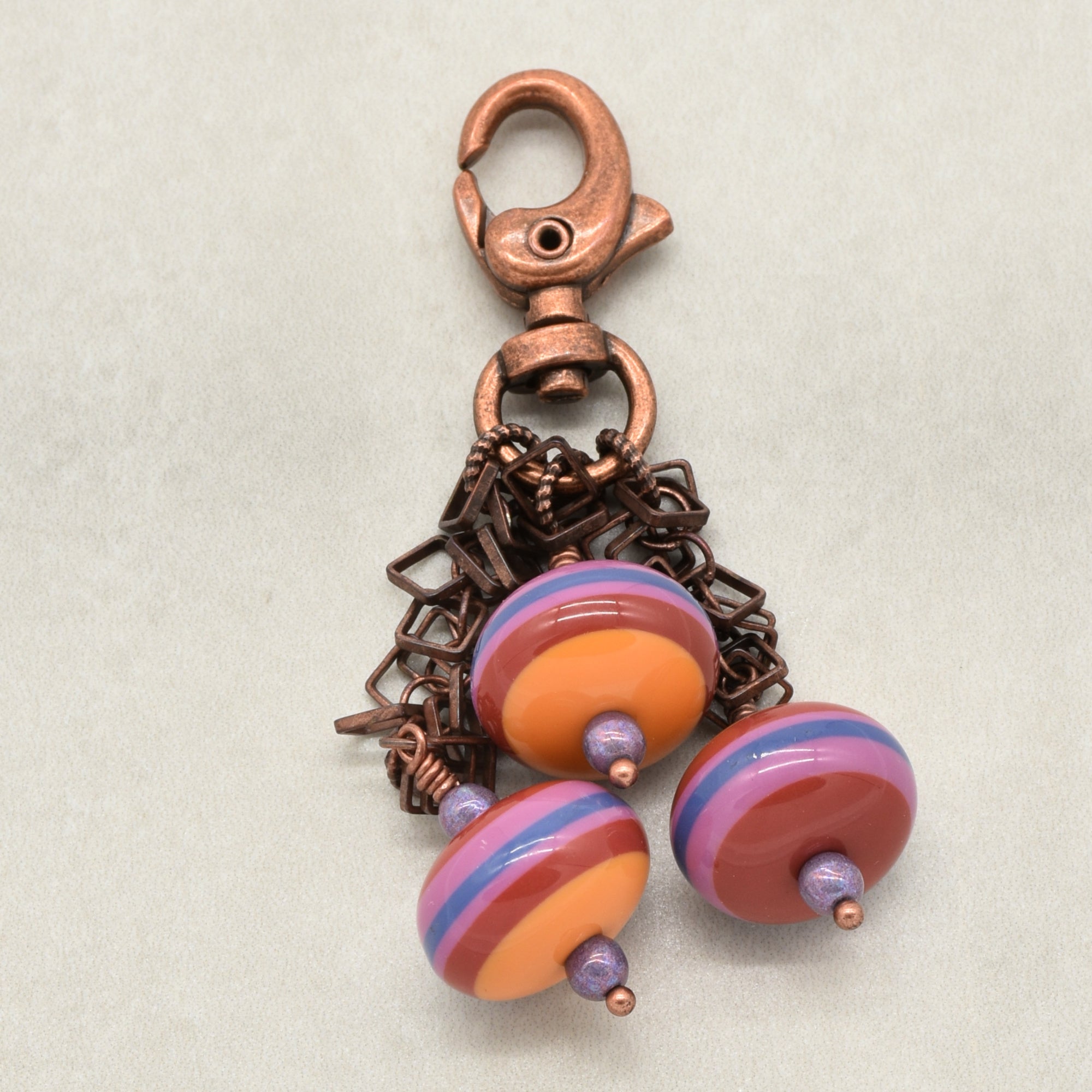 A handmade copper-toned lobster clasp keychain or bag charm featuring three colorful striped vintage Lucite beads in orange, purple, and blue, set against a neutral light gray background - made with love in Colorado by Ruby Acorn.
