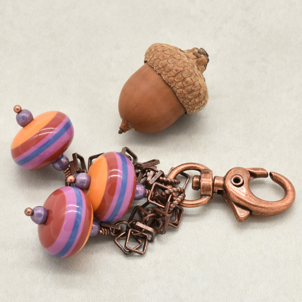 A copper-toned keychain or bag charm with three vintage Lucite striped beads in orange, pink, and purple, displayed next to an acorn on a light gray background -made with love in Colorado by Ruby Acorn.