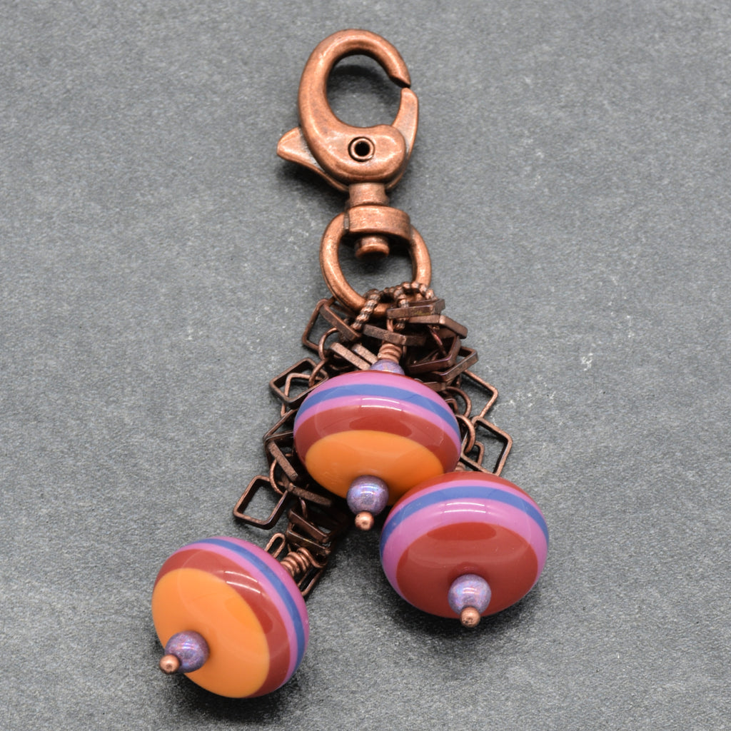 A handmade copper keychain featuring a lobster clasp and three dangling vintage Lucite beads with orange, purple, and blue striped patterns, set against a textured gray background - made with love in Colorado by Ruby Acorn.