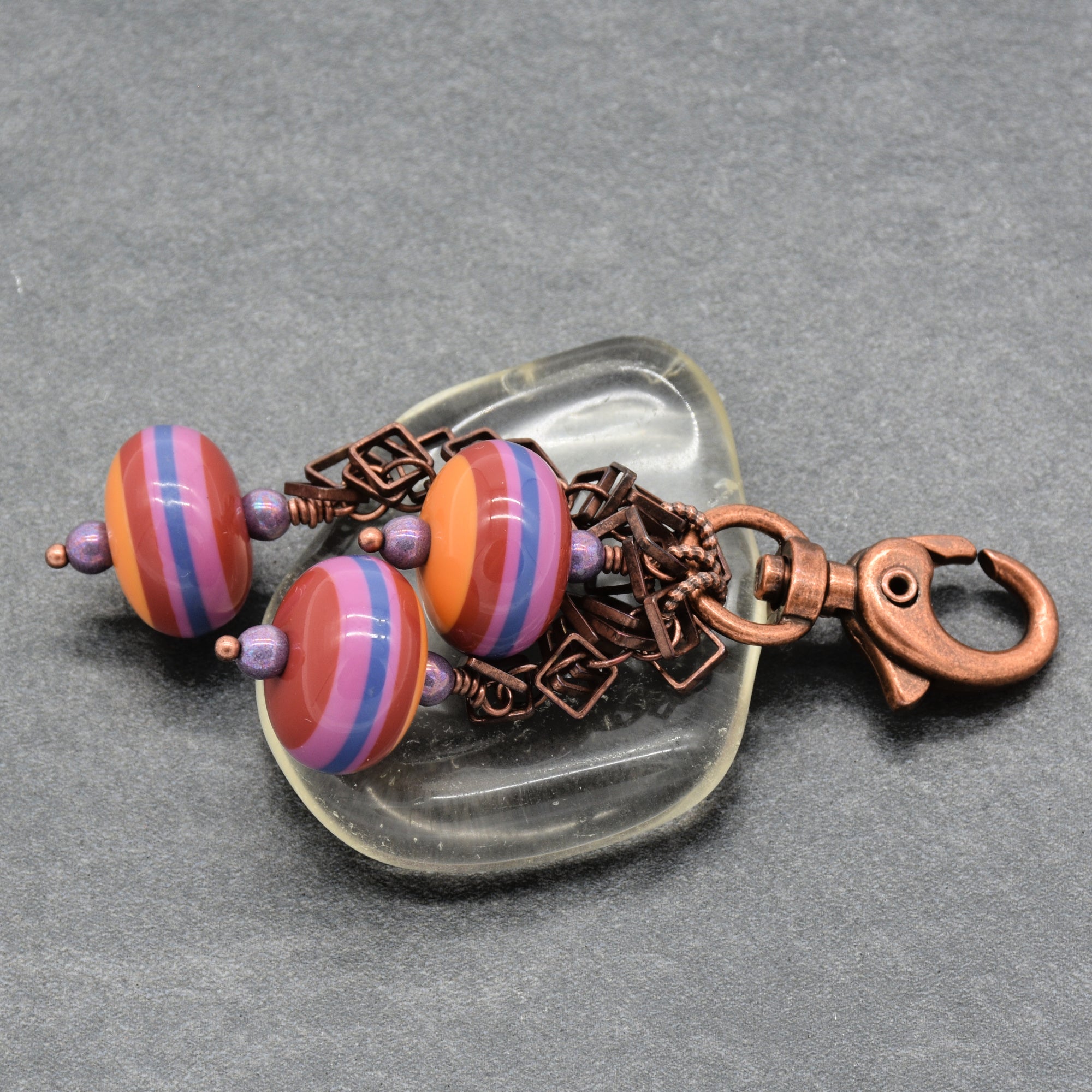A pair of handmade charm with striped vintage Lucite beads in orange, pink, and purple, finished with a copper lobster clasp, displayed on a clear glass dish over a gray textured background - made with love in Colorado by Ruby Acorn.