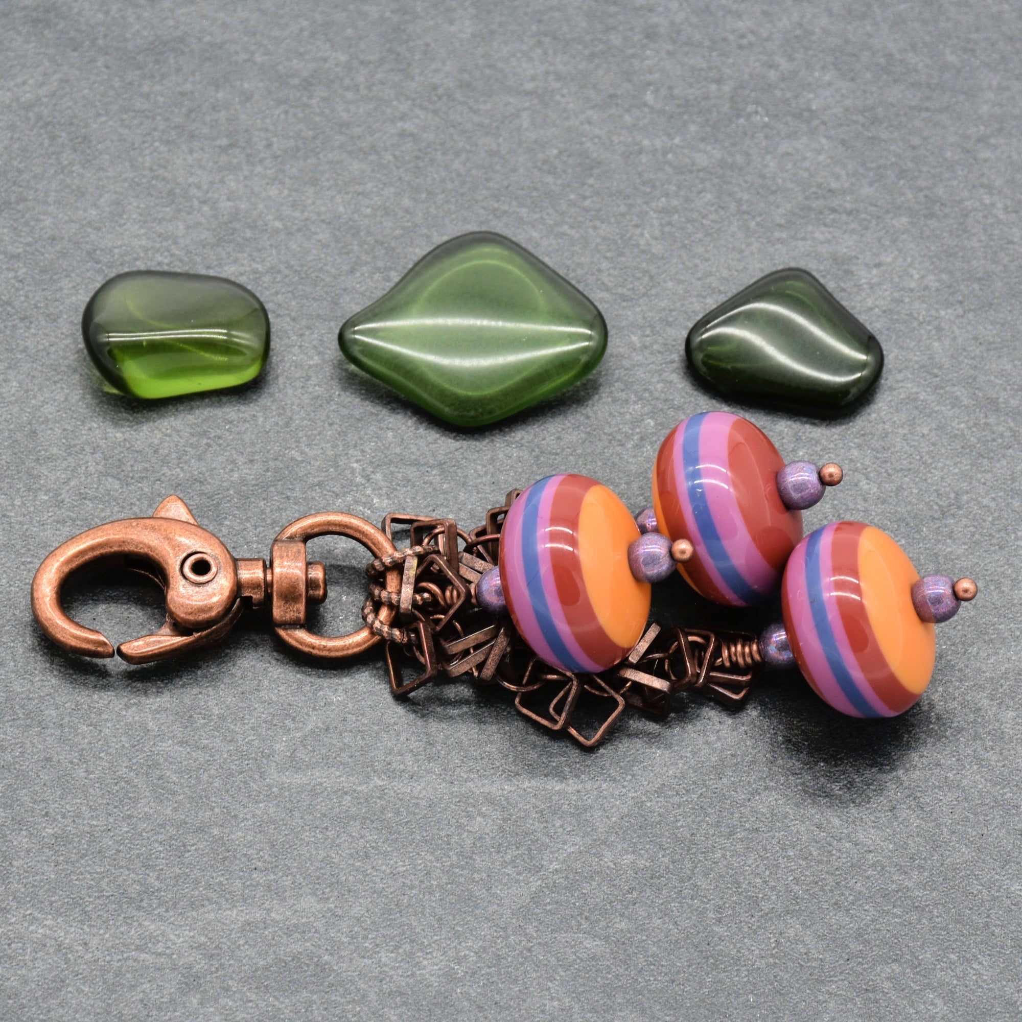 A keychain charm set on a gray textured background, three vintage Lucite striped beads in orange, pink, and blue with purple spacers, and a copper clasp and chain - made with love in Colorado by Ruby Acorn.