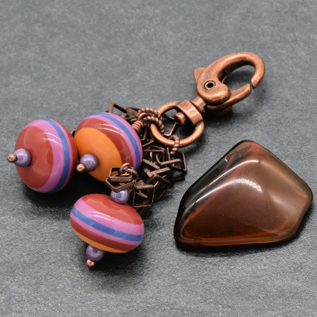 A handmade jewelry charm set featuring a copper-toned lobster clasp, three colorful striped vintage Lucite beads in orange, pink, and blue, displayed on a dark gray textured background - made with love in Colorado by Ruby Acorn.