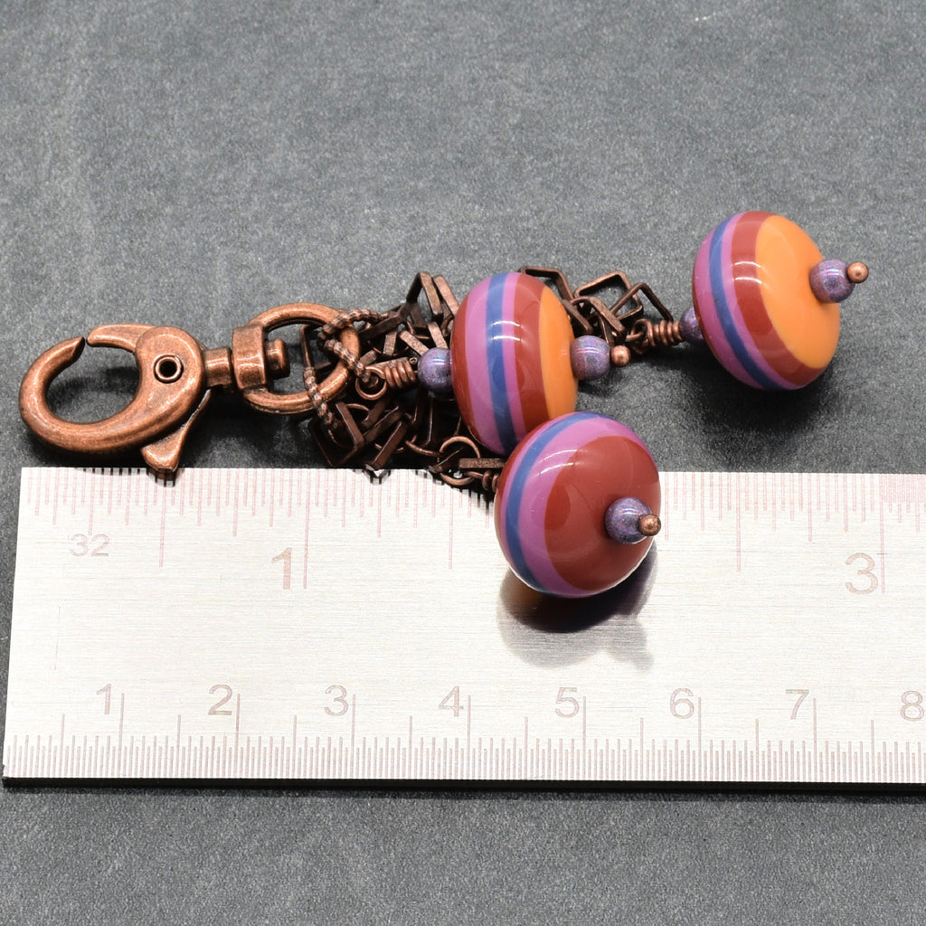 A handmade keychain or bag charm featuring three striped vintage Lucite beads in orange, purple, and blue, attached to a copper-toned metal clasp and chain, shown next to a ruler for scale on a dark gray background - made with love by Ruby Acorn in Colorado.