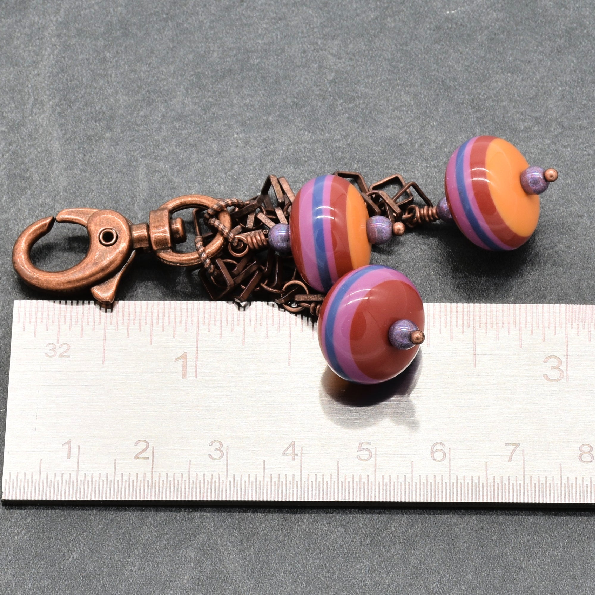 A handmade keychain or bag charm featuring three striped vintage Lucite beads in orange, purple, and blue, attached to a copper-toned metal clasp and chain, shown next to a ruler for scale on a dark gray background - made with love by Ruby Acorn in Colorado.