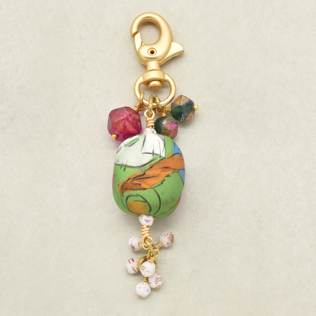 A gold-tone lobster clasp keychain charm featuring a colorful hand-painted green and orange clay bead, small pink and green gemstones, and dangling white beads with pink speckles, set against a neutral light gray background - made with love in Colorado by Ruby Acorn.