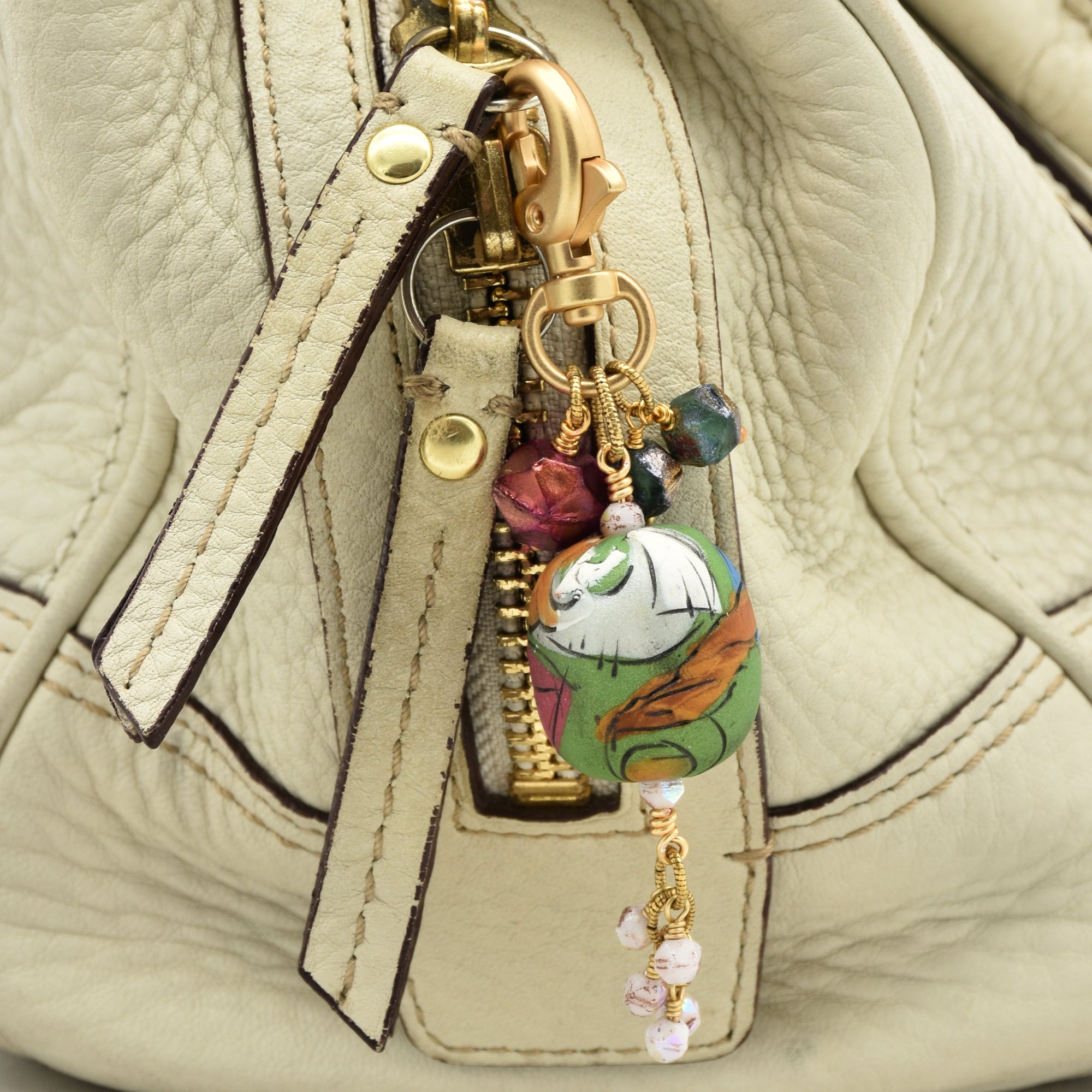 A close-up of a beige textured leather handbag featuring a decorative gold-tone keychain charm with colorful beads and a round painted character bead - made with love in Colorado by Ruby Acorn.