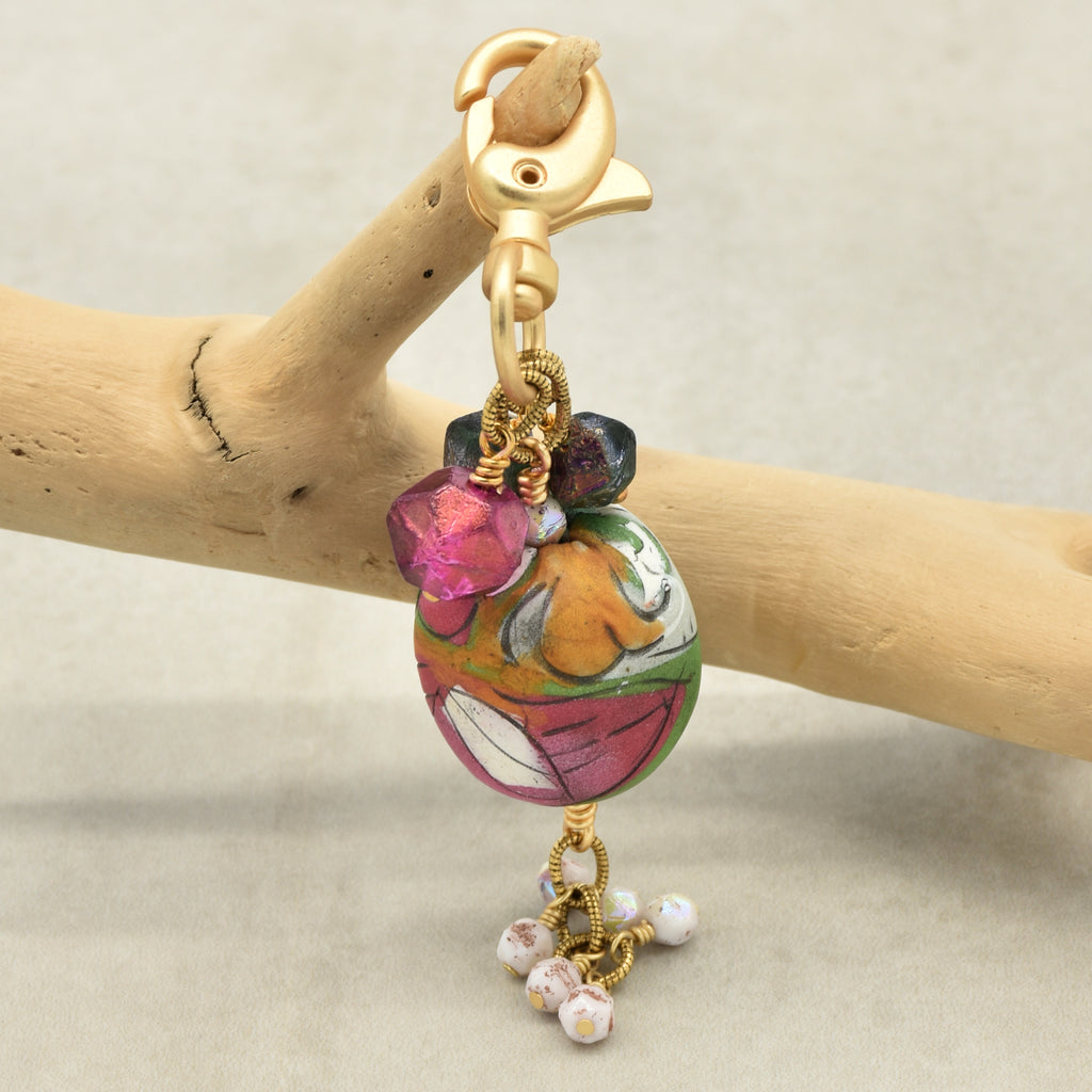 A gold-tone lobster clasp keychain or bag charm featuring a colorful hand-painted glass bead with pink, green, orange, and white patterns, adorned with small dangling beads, displayed on a light wood branch against a neutral beige background - made with love in Colorado by Ruby Acorn.