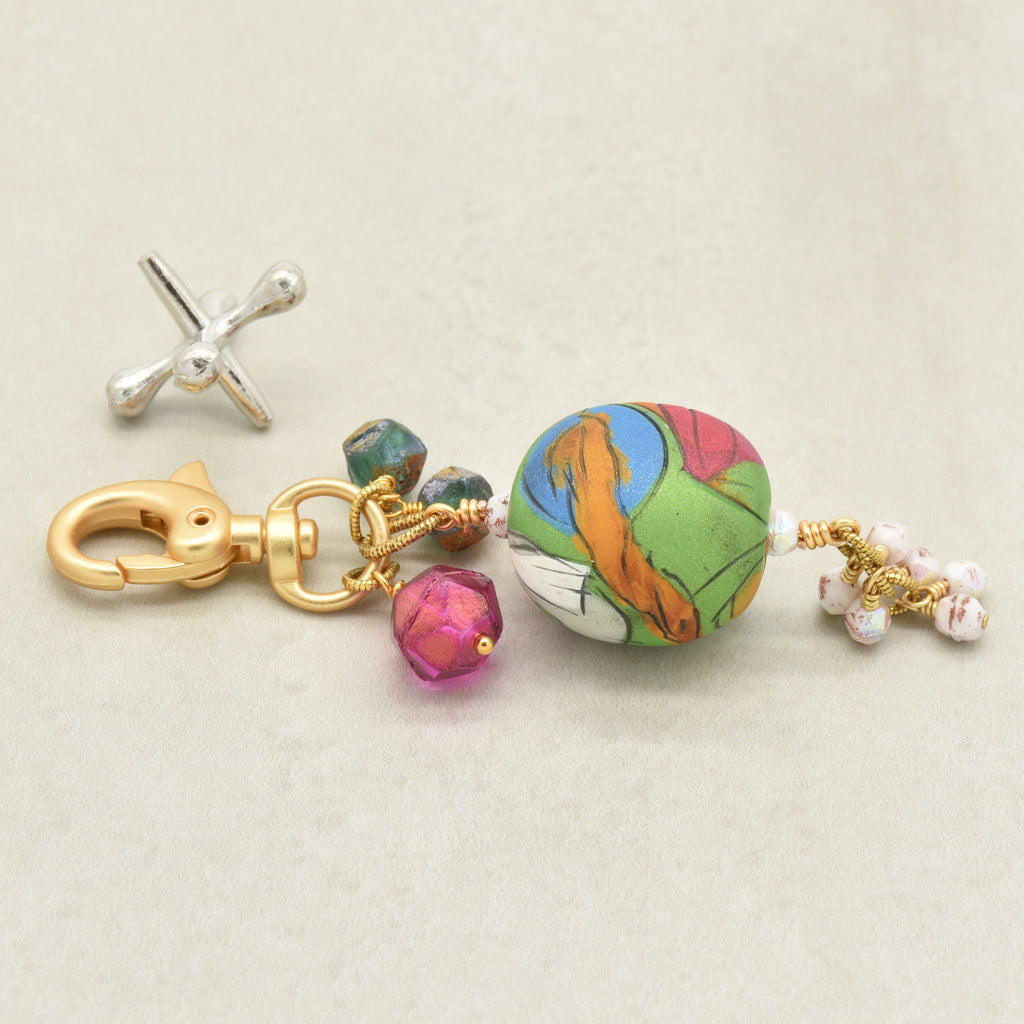 A handmade keychain featuring a colorful artisan polymer clay marbled bead with green, orange, pink, and blue patterns, gold-plated hardware, faceted magenta and sea green beads, and small white pearl-like beads on a light beige background - made with love in Colorado by Ruby Acorn.