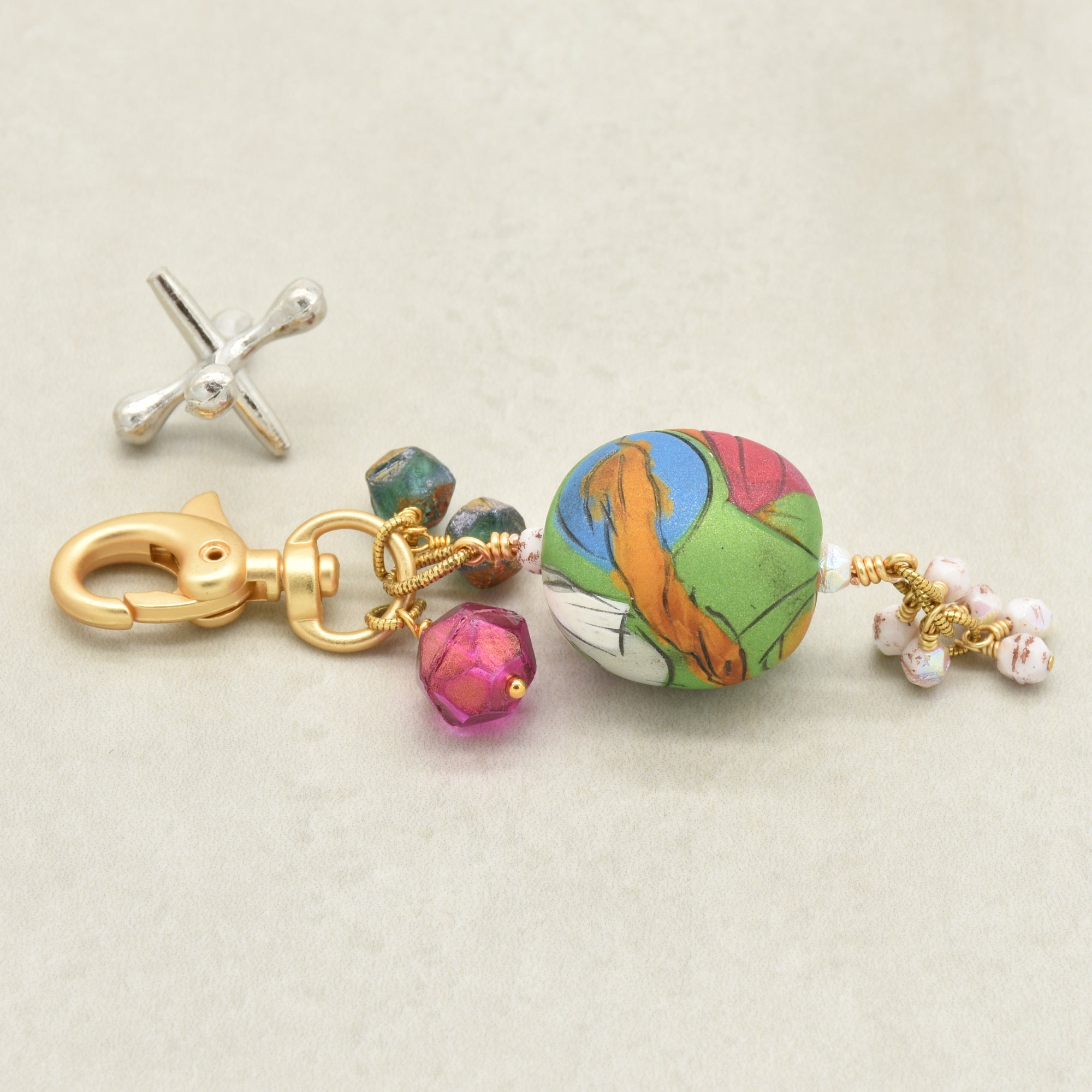 A handmade keychain featuring a colorful artisan polymer clay marbled bead with green, orange, pink, and blue patterns, gold-plated hardware, faceted magenta and sea green beads, and small white pearl-like beads on a light beige background - made with love in Colorado by Ruby Acorn.