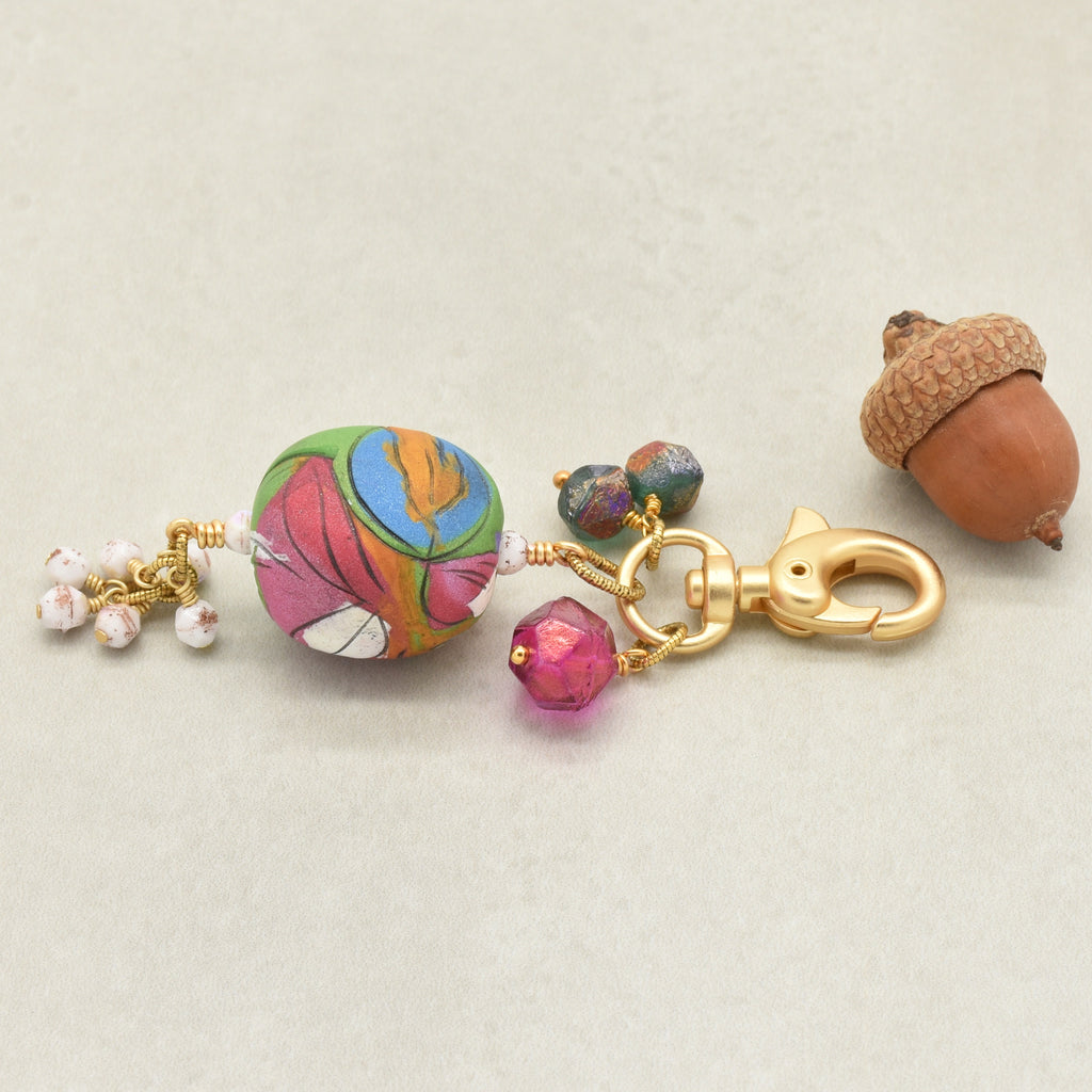 A handmade keychain charm featuring a colorful artisan polymer clay marbled bead, a gold lobster clasp, a faceted pink bead, and small white pearl-like beads, styled with a brown acorn on a light beige background - made with love in Colorado by Ruby Acorn.