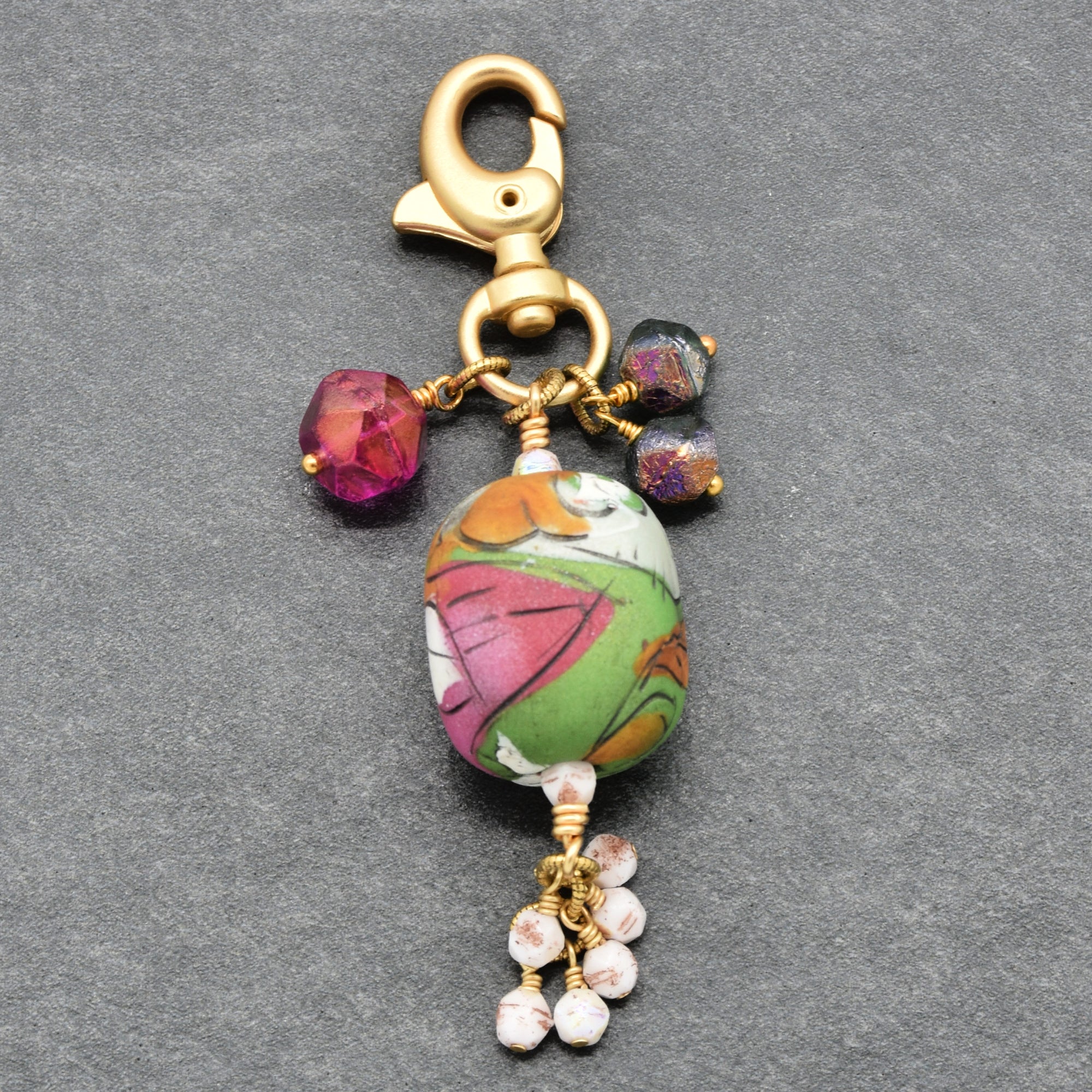 A handmade keychain charm featuring an artisan polymer clay bead with a pink, green, orange, and white marbled pattern, accented by small faceted beads in magenta, purple, and white, all attached to a gold-tone lobster clasp on a textured gray background - made with love by Ruby Acorn in Colorado.