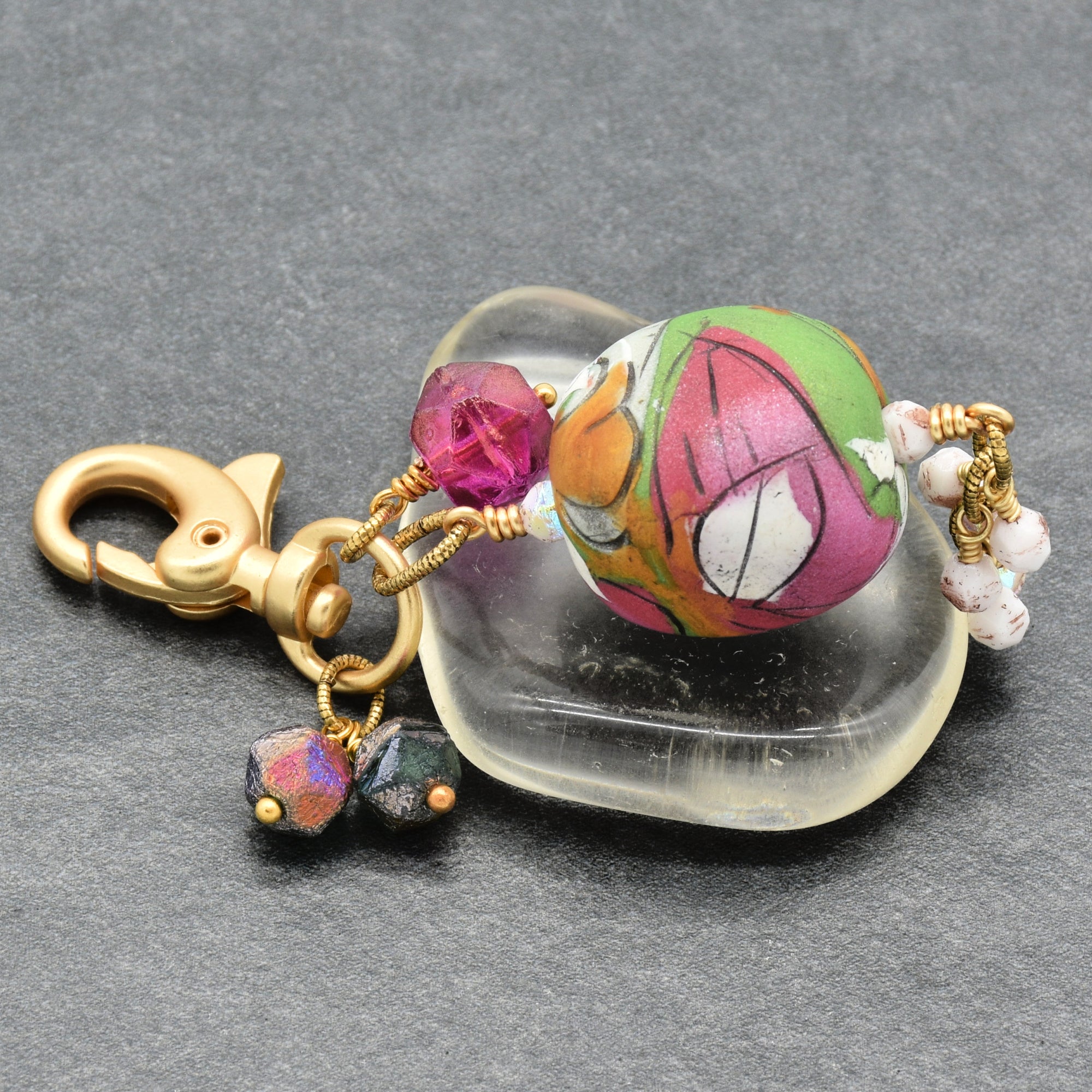 A handmade beaded keychain charm with a colorful floral pattern, gold clasp, and dangling beads, displayed on a clear stone over a dark gray textured background - made with love in Colorado by Ruby Acorn.