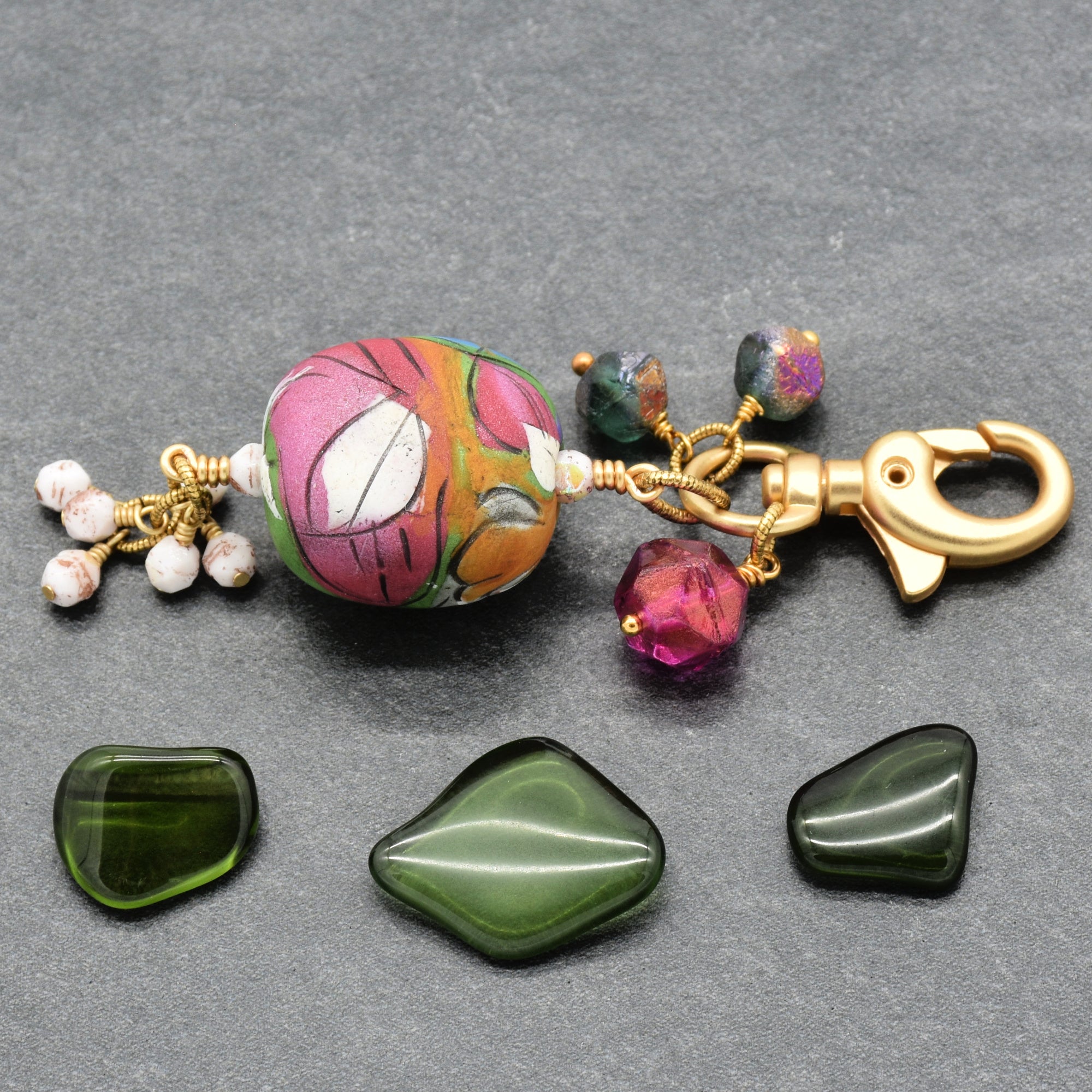 A handmade keychain or bag charm with an artisan polymer clay marbled pink, orange, and white bead, gold-tone hardware, and small green and fuchsia Czech glass beads, displayed on a gray background with three green stone beads below - made with love in Colorado by Ruby Acorn.
