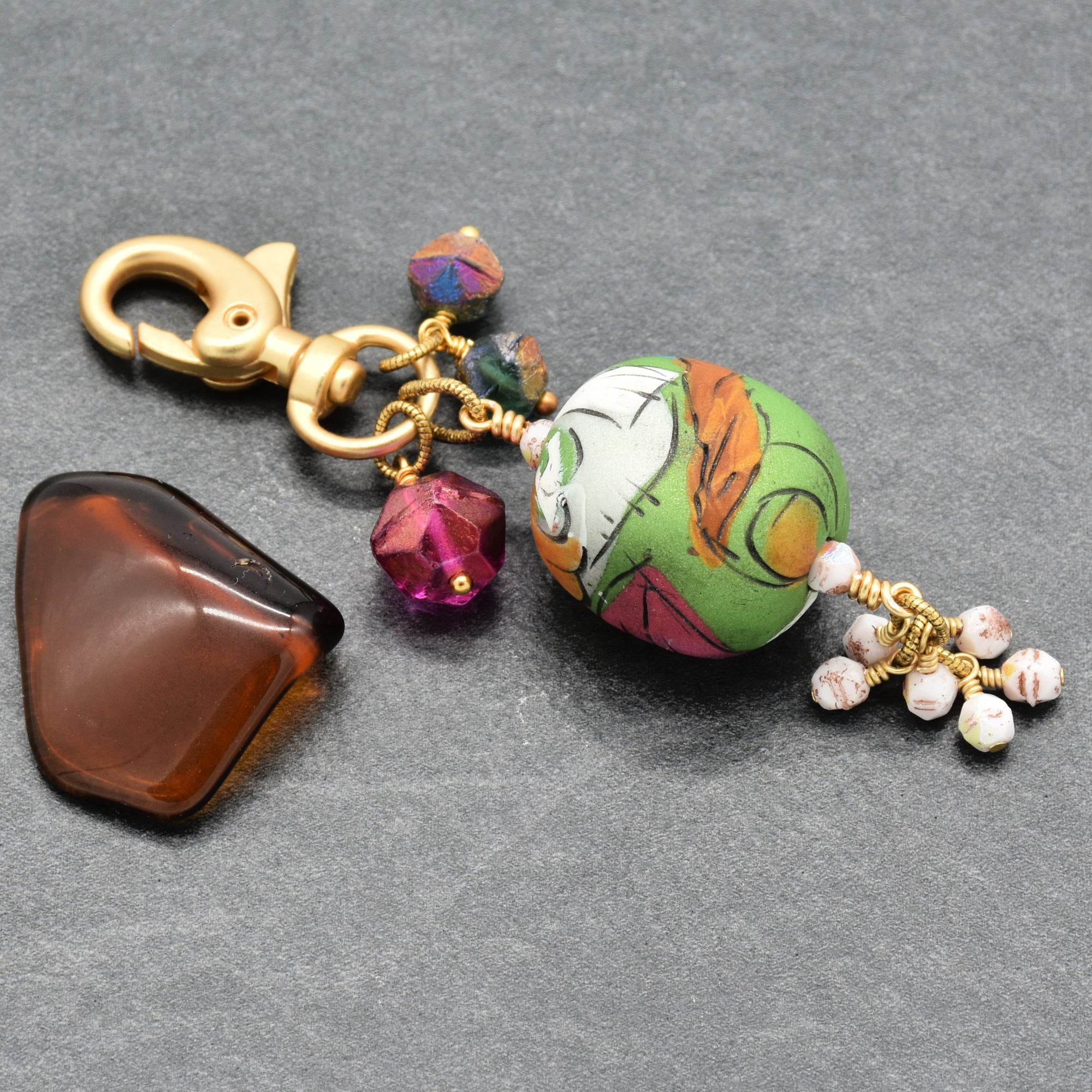 A handmade keychain charm featuring an artisan polymer clay large green and white patterned focal, a faceted magenta, sea green and white Czech glass beads, and a gold clasp, displayed on a dark gray textured background - made with love in Colorado by Ruby Acorn.