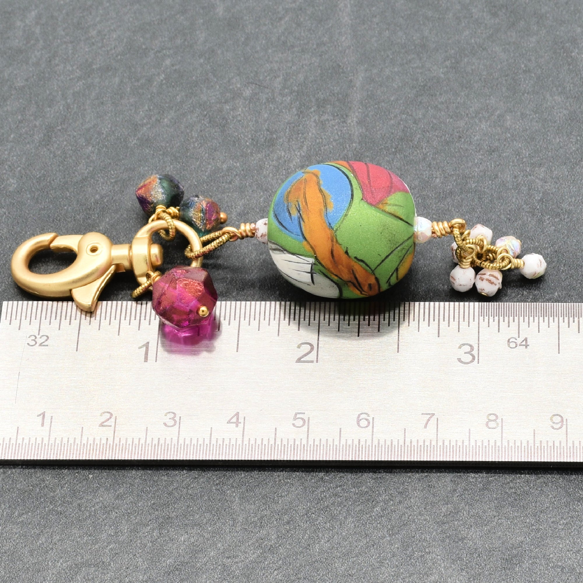 A handmade keychain or bag charm featuring a colorful marbled bead with gold-toned hardware, a lobster clasp, and a tassel of small white beads, shown next to a metal ruler for scale on a dark gray background - made with love in Colorado by Ruby Acorn.