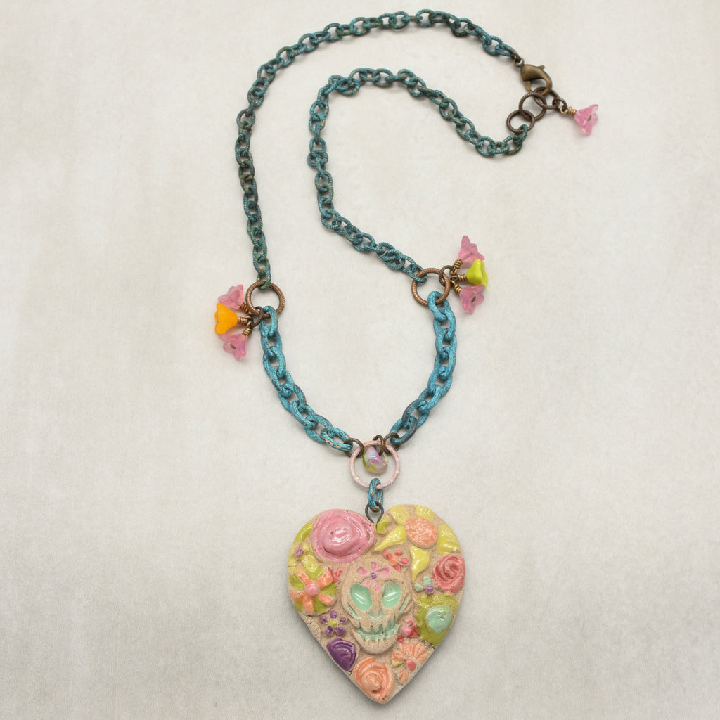 A handmade verdigris chain necklace with a Dia de los Muertos heart-shaped sugar skull pendant decorated with pink, yellow, and green floral details, shown on a textured off-white background - made with love in Colorado by Ruby Acorn.