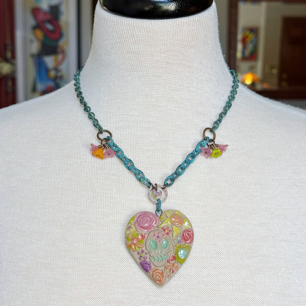 A colorful heart-shaped Dia de los Muertos pendant necklace displayed on a white fabric mannequin bust. The necklace features a verdigris chain with small dangling bellflower charms and a large heart pendant decorated with a pastel sugar skull and floral patterns - made with love in Colorado by Ruby Acorn.