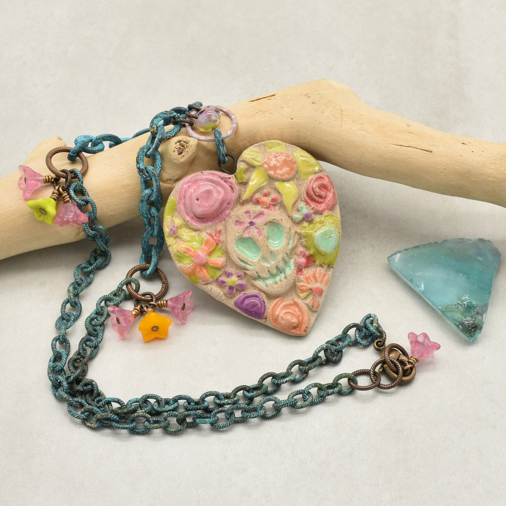 A handmade Dia de los Muertos heart-shaped sugar skull pendant necklace with a verdigris chain, displayed on a light wood driftwood piece with a small blue stone accent on a neutral background - made with love in Colorado by Ruby Acorn.