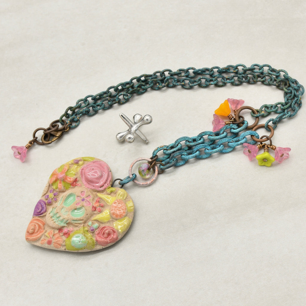 A handmade verdigris chain necklace with a large, colorful heart-shaped Dia de los Muertos sugar skull pendant, and small pink, green and orange flower charms, on a light beige background - made with love in Colorado by Ruby Acorn.