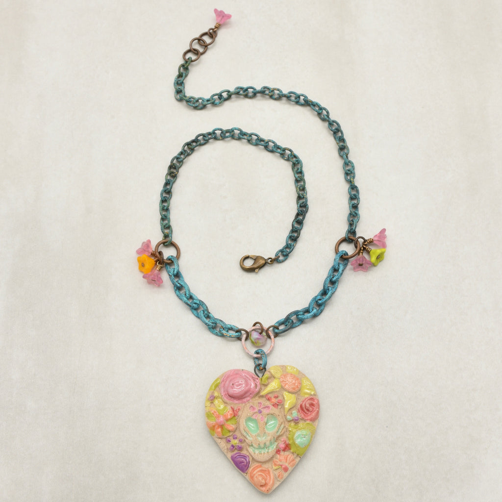 A handmade verdigris chain necklace with a colorful Dia de los Muertos sugar skull heart pendant decorated with pink, yellow, and green floral details, shown on a light beige textured background - made with love in Colorado by Ruby Acorn.