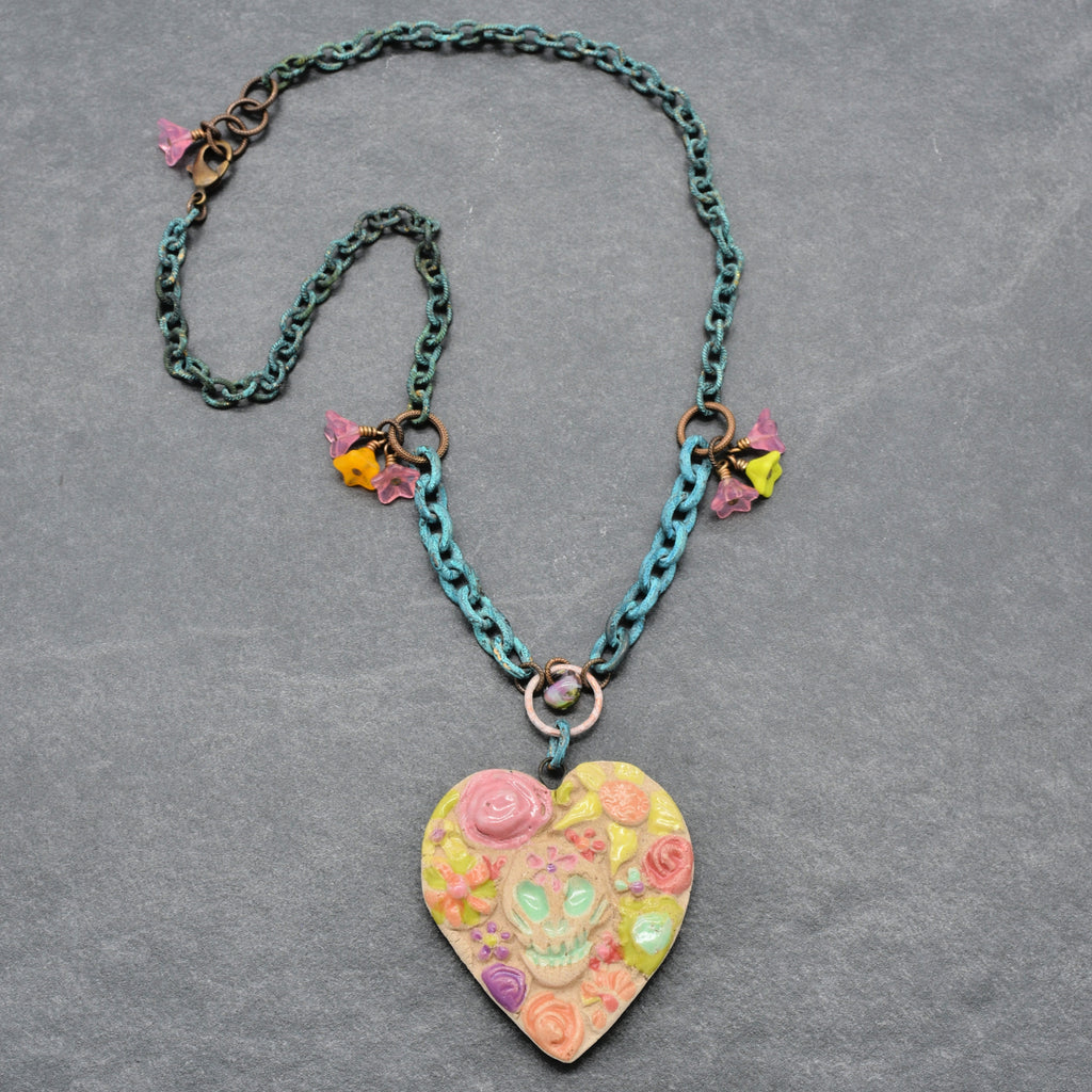 A handmade verdigris chain necklace with a Day of the Dead heart-shaped pendant featuring a colorful sugar skull and floral design, displayed on a textured gray background - made with love in Colorado by Ruby Acorn.