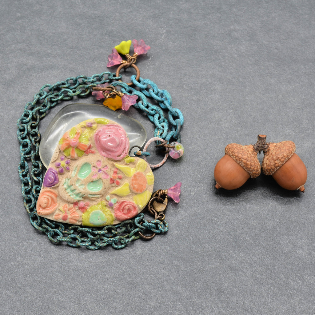 A handmade Day of the Dead pendant necklace featuring a colorful sugar skull and floral heart charm, layered with verdigris chain, displayed next to a pair of acorns on a textured gray background - made with love in Colorado by Ruby Acorn.