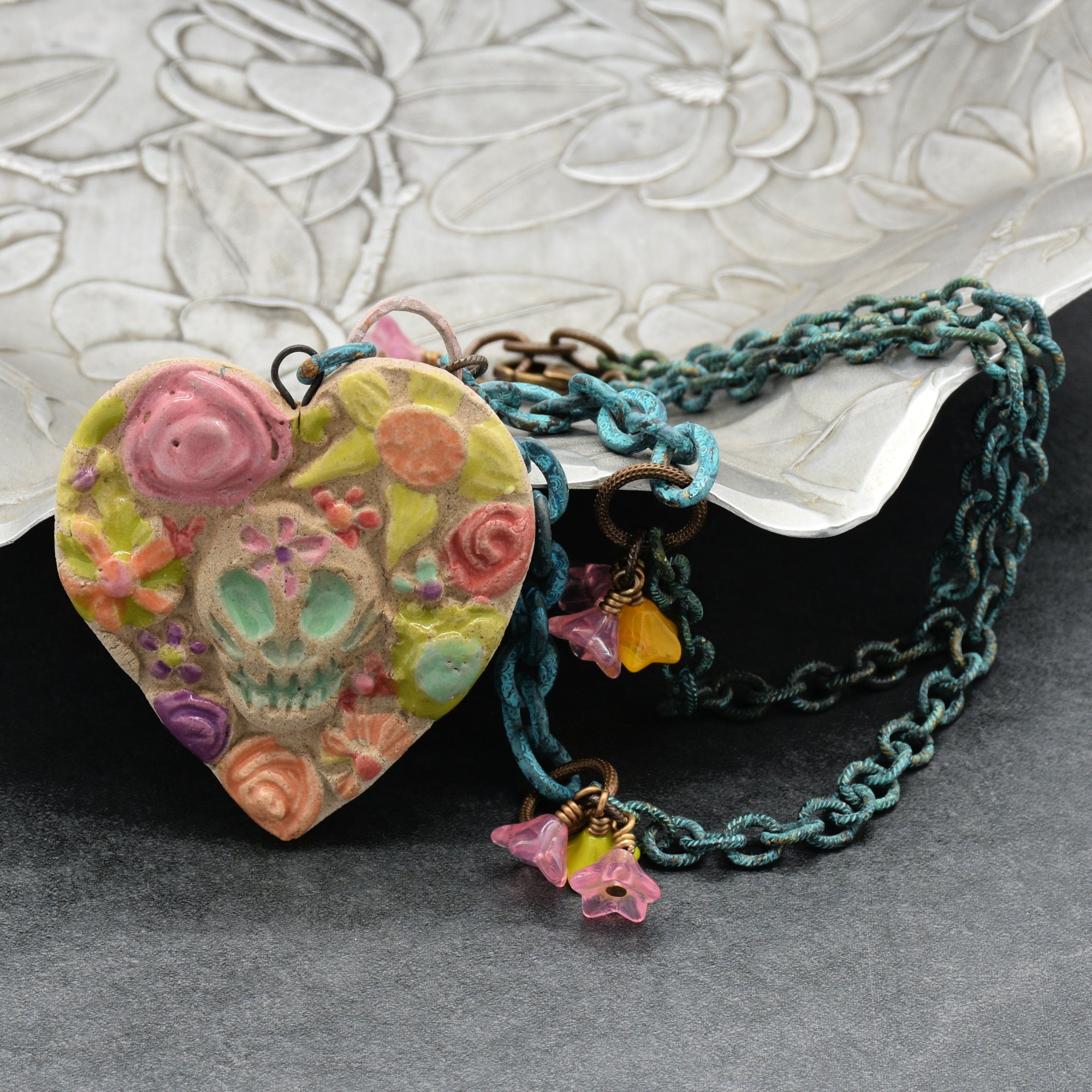 A handmade heart-shaped pendant necklace with a colorful Day of the Dead sugar skull and floral design, attached to a verdigris chain with small dangling flower charms, displayed on a textured silver surface - made with love in Colorado by Ruby Acorn.