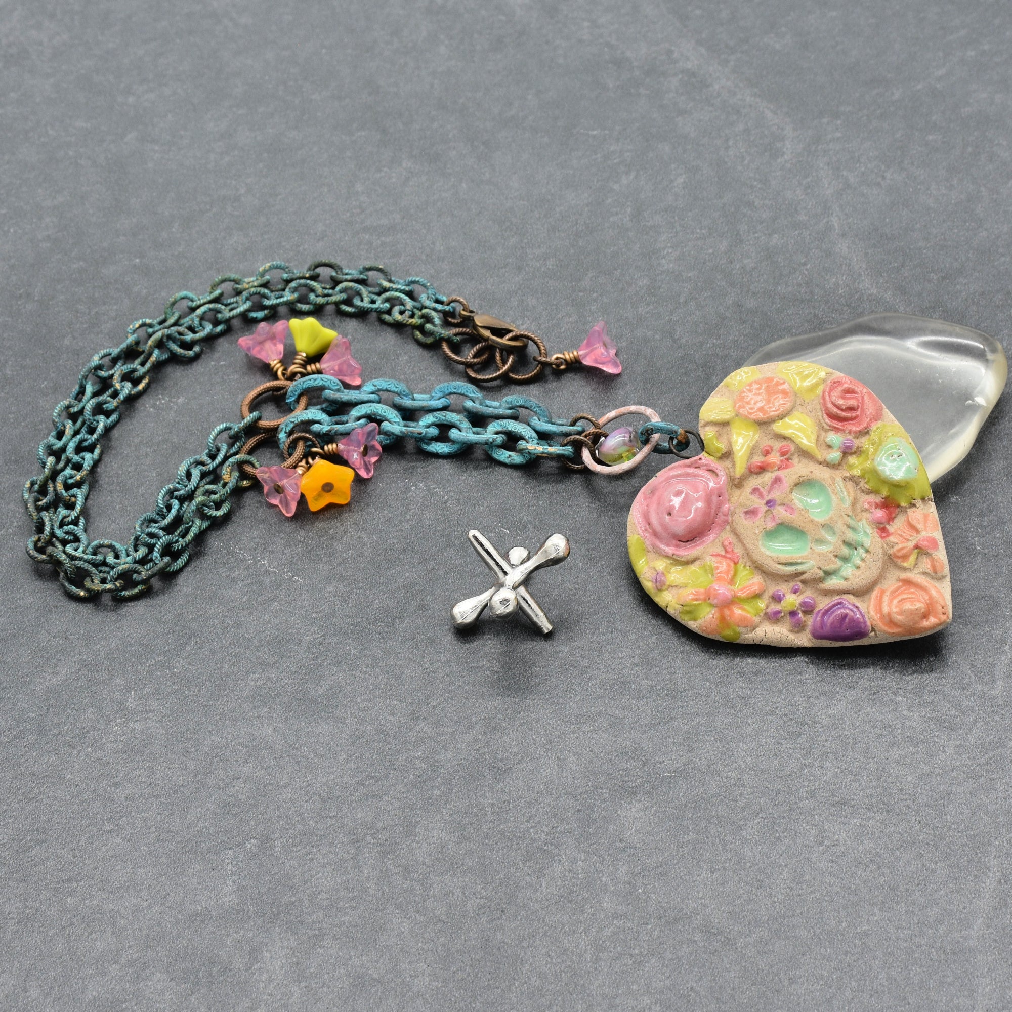 A handmade verdigris chain necklace with a colorful floral Day of the Dead heart pendant, displayed on a gray textured background - made with love in Colorado by Ruby Acorn.