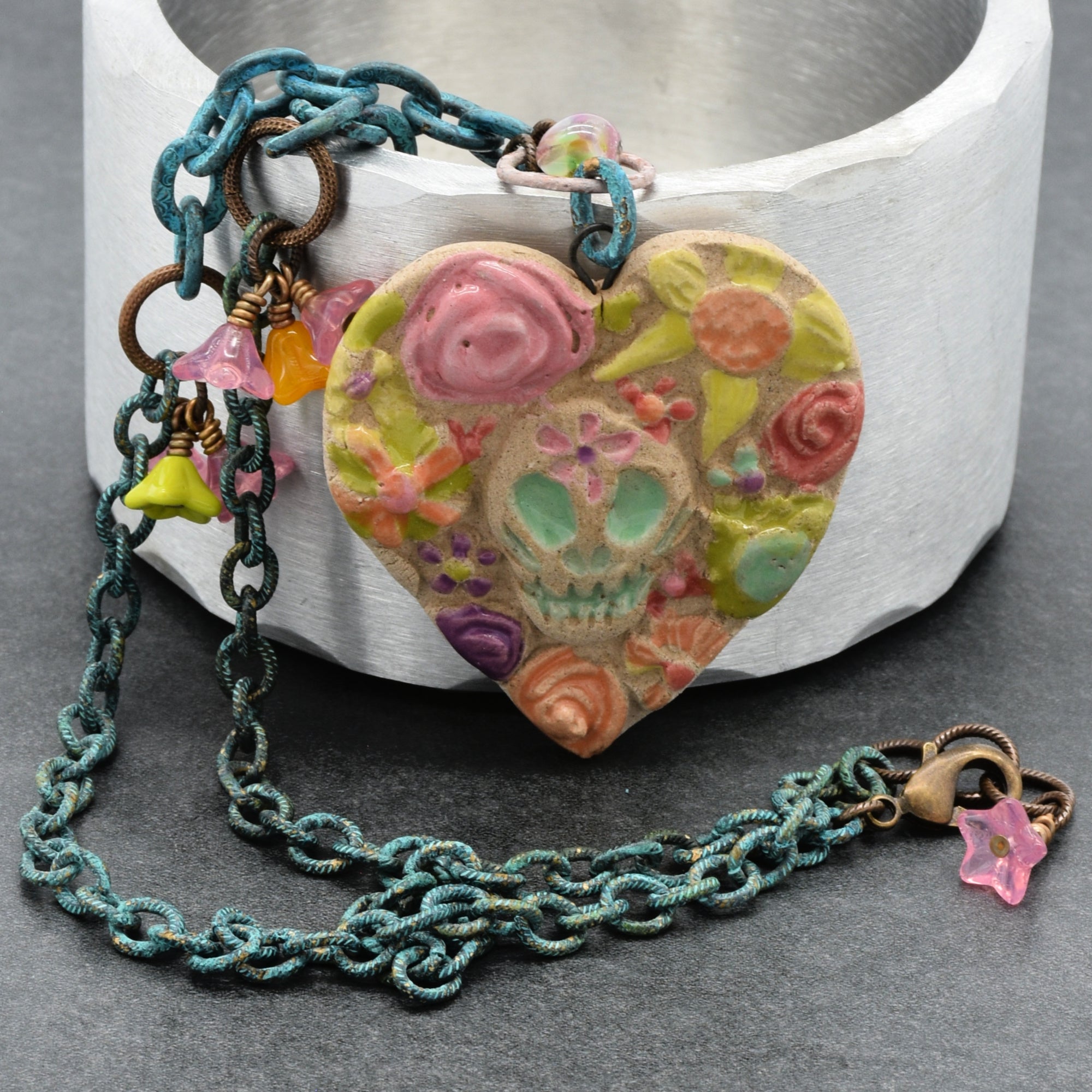 A heart-shaped Day of the Dead pendant necklace with a colorful floral and sugar skull design, hanging from a verdigris chain with antique brass finish links and small dangling bellflower charms in pink, lime green and orange, displayed in front of a silver cylindrical container on a gray surface - made with love in Colorado by Ruby Acorn.