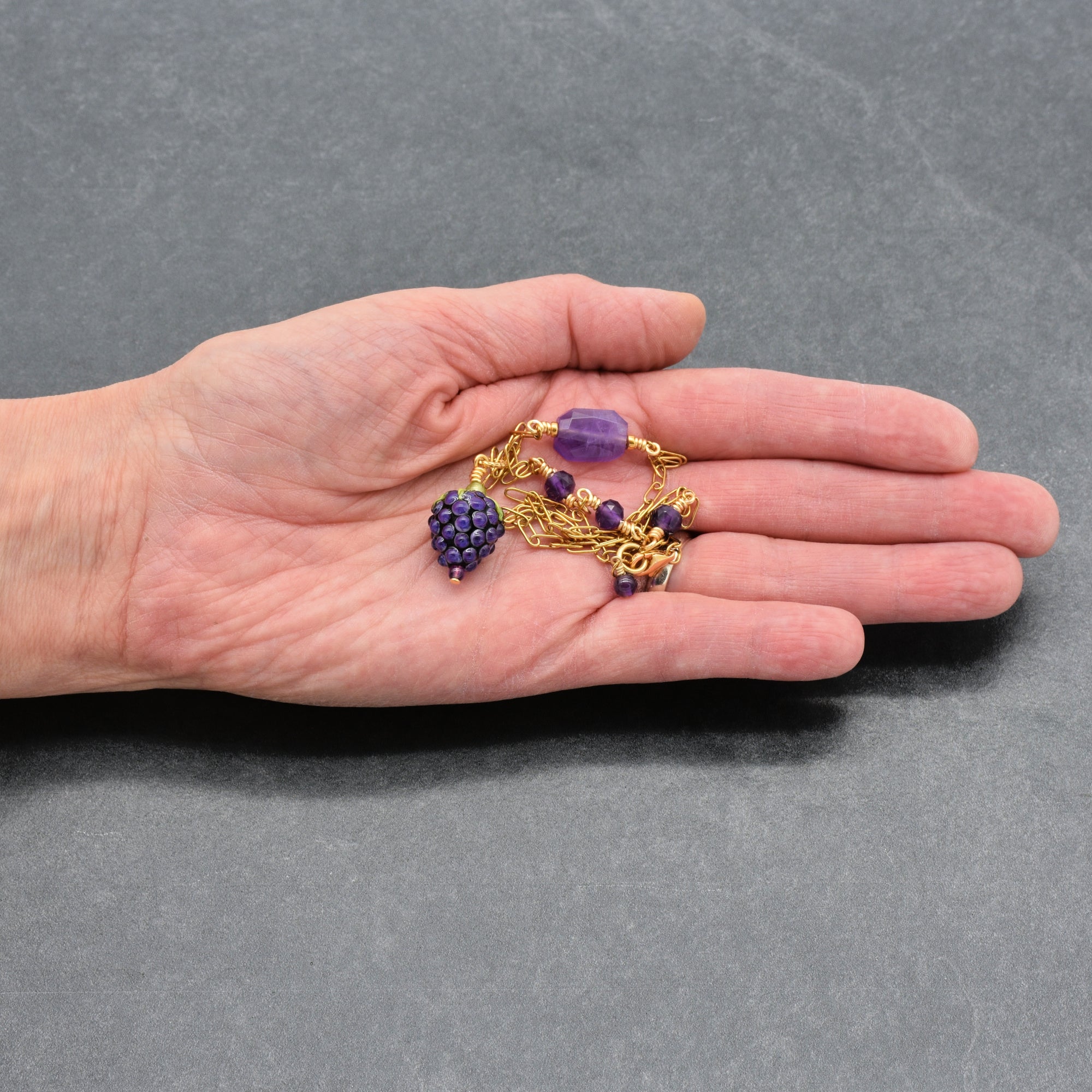 Artist's hand holding asymmetrical gold chain necklace with artisan lampwork glass blackberry focal and amethyst on a gray background - handmade with love by Ruby Acorn