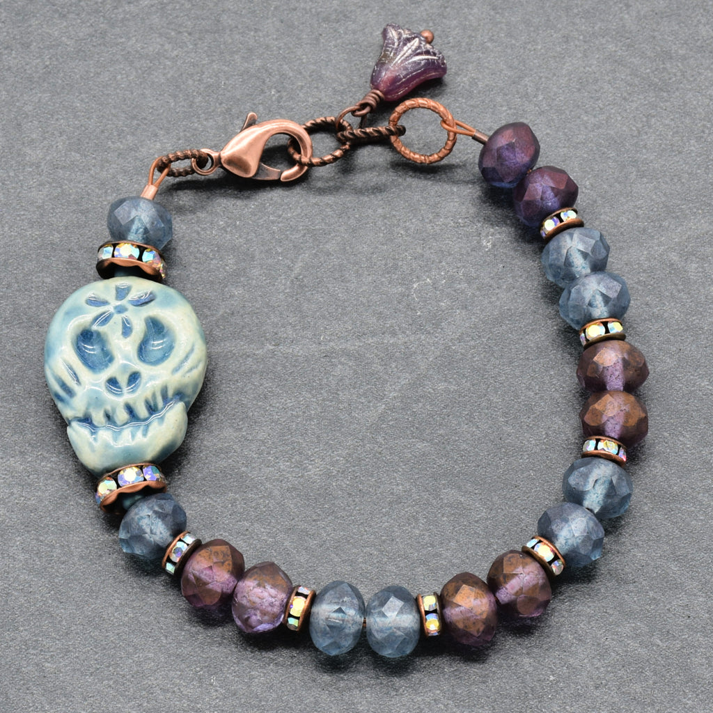 A beaded bracelet featuring a blue Dia de los Muertos sugar skull, faceted beads in shades of blue & purple, bronze Rhinestones, and a copper-toned clasp on a textured gray background - made with love in Colorado by Ruby Acorn.