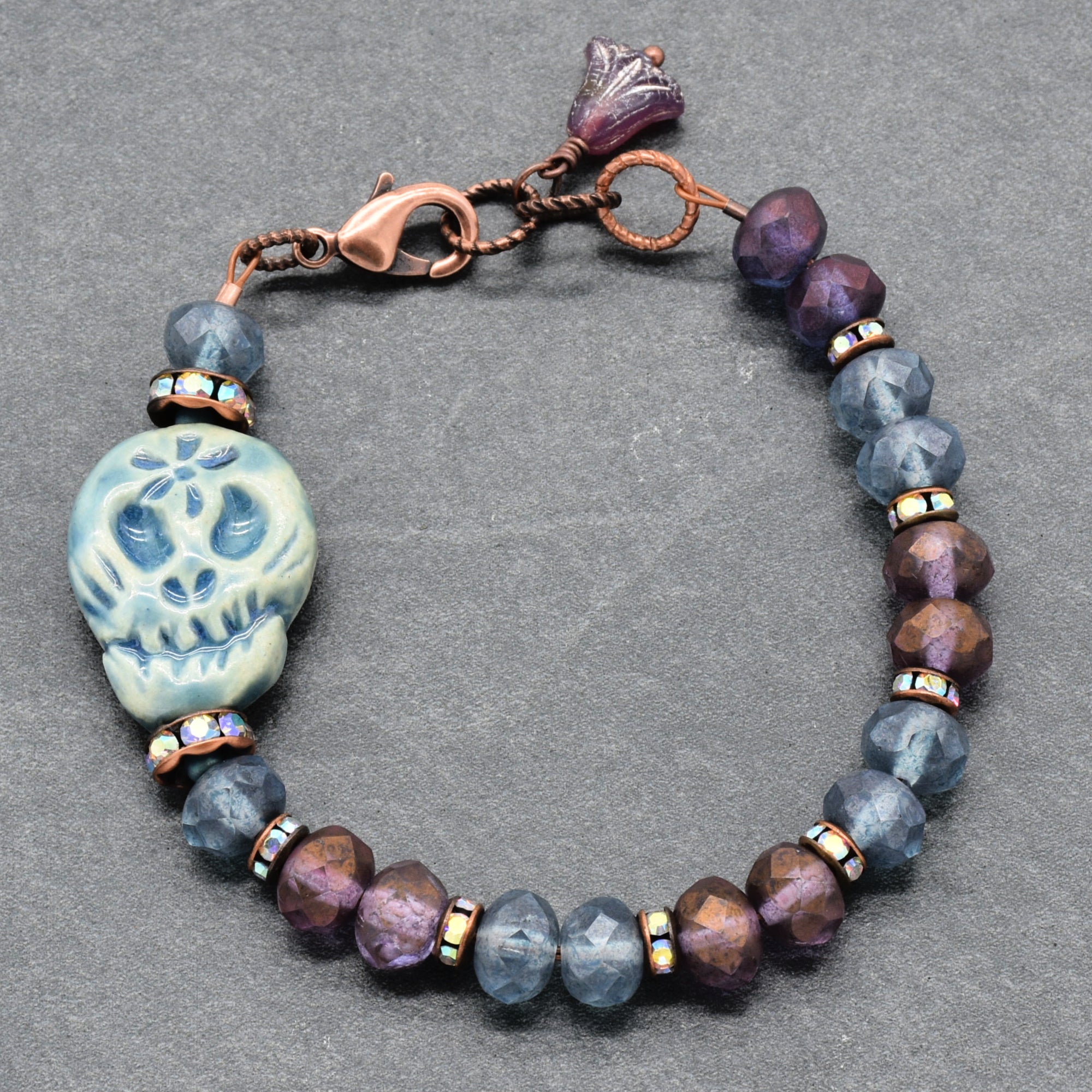 A beaded bracelet featuring a blue Dia de los Muertos sugar skull, faceted beads in shades of blue & purple, bronze Rhinestones, and a copper-toned clasp on a textured gray background - made with love in Colorado by Ruby Acorn.