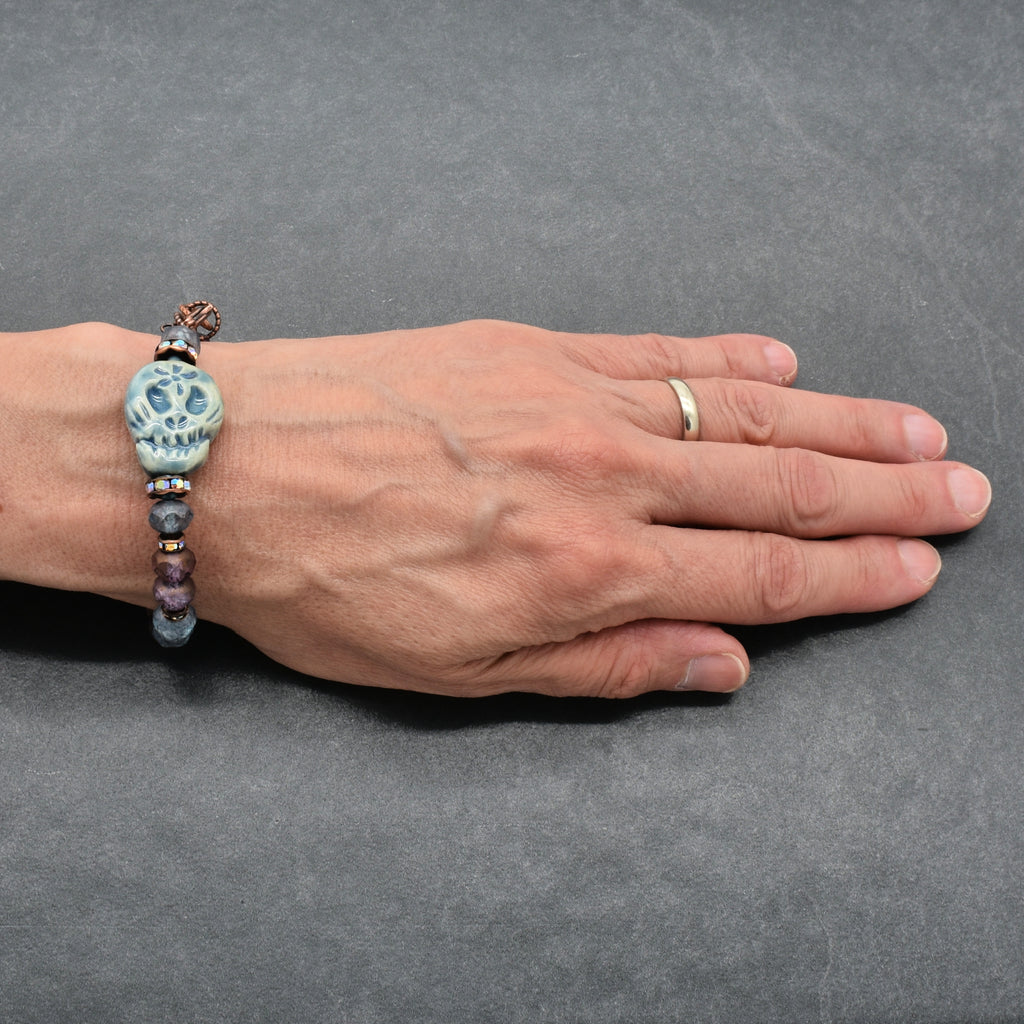 A beaded bracelet with a blue Day of the Dead sugar skull and dark beads worn on a person's wrist, shown against a dark gray textured background - made with love in Colorado by Ruby Acorn.