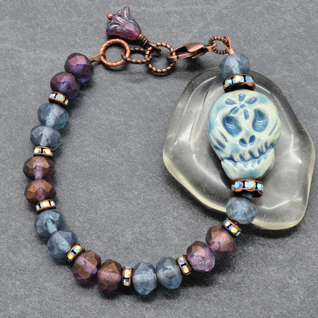 A handmade beaded bracelet with faceted blue and purple beads, a copper-toned clasp, and a central blue Dia de los Muertos sugar skull, displayed on a gray textured background next to a clear stone - made with love in Colorado by Ruby Acorn.