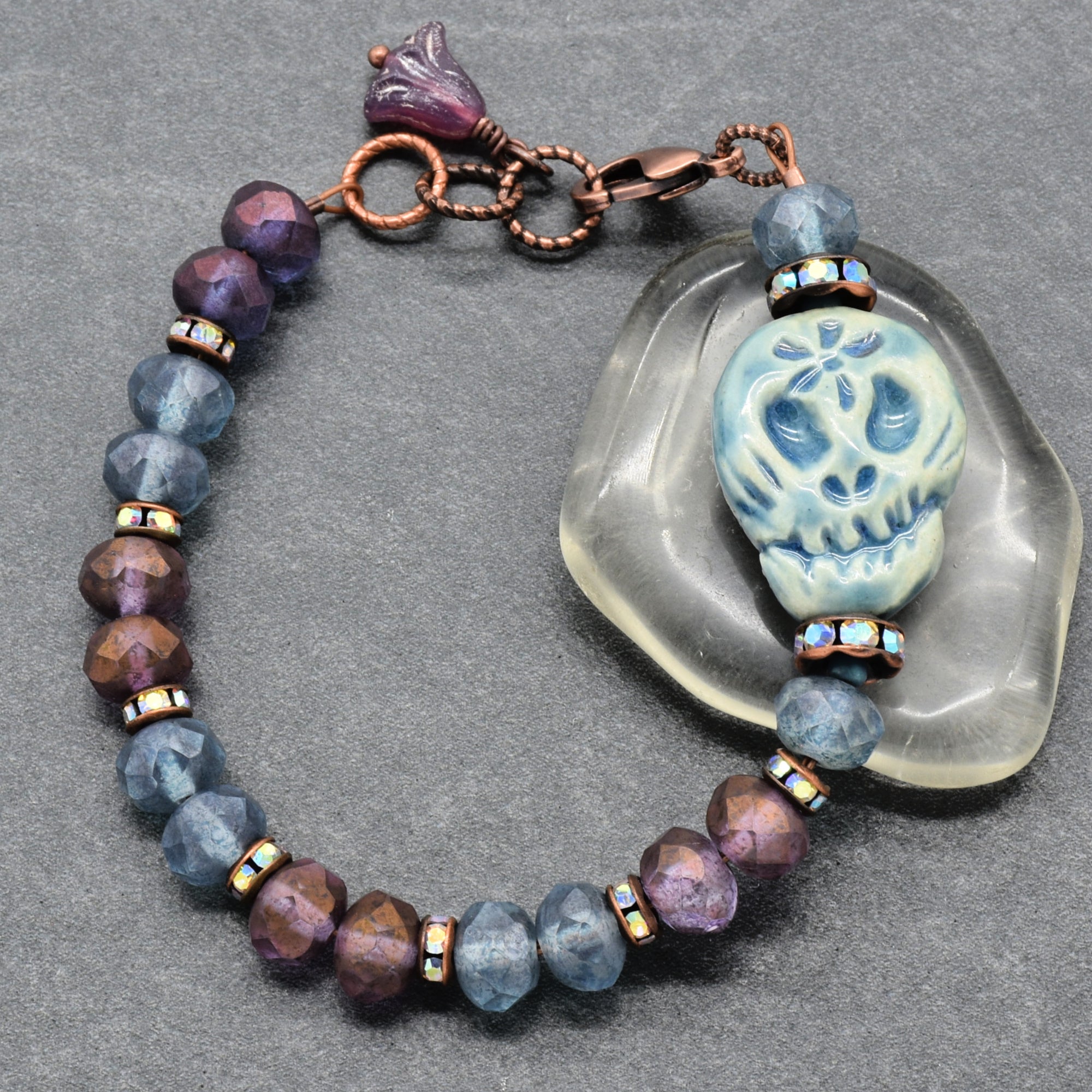 A handmade beaded bracelet with faceted blue and purple beads, a copper-toned clasp, and a central blue Dia de los Muertos sugar skull, displayed on a gray textured background next to a clear stone - made with love in Colorado by Ruby Acorn.