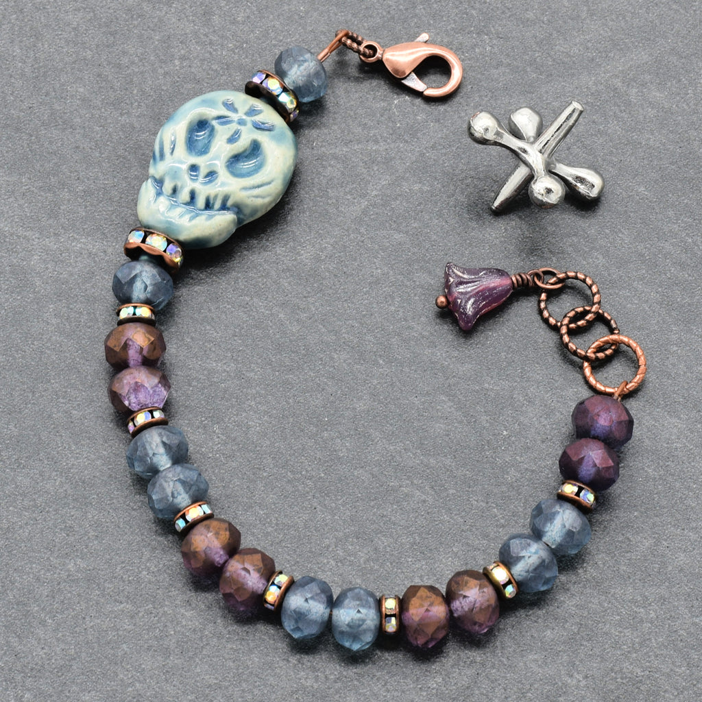 A beaded bracelet with faceted blue-gray and purple beads, a Dia de Los Muertos sugar skull bead, a copper clasp, and a purple flower charm, displayed on a dark gray textured background - made with love in Colorado by Ruby Acorn.