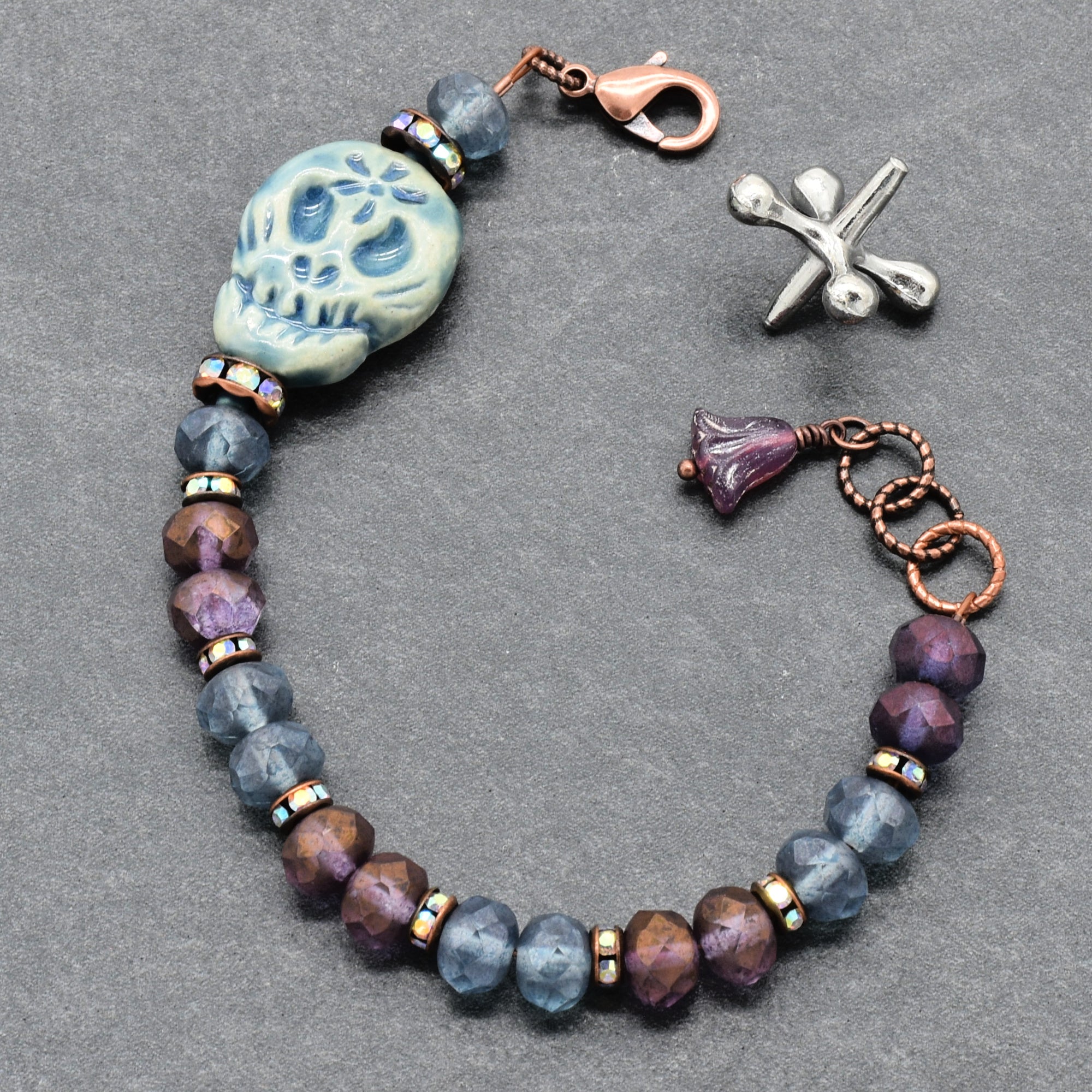A beaded bracelet with faceted blue-gray and purple beads, a Dia de Los Muertos sugar skull bead, a copper clasp, and a purple flower charm, displayed on a dark gray textured background - made with love in Colorado by Ruby Acorn.