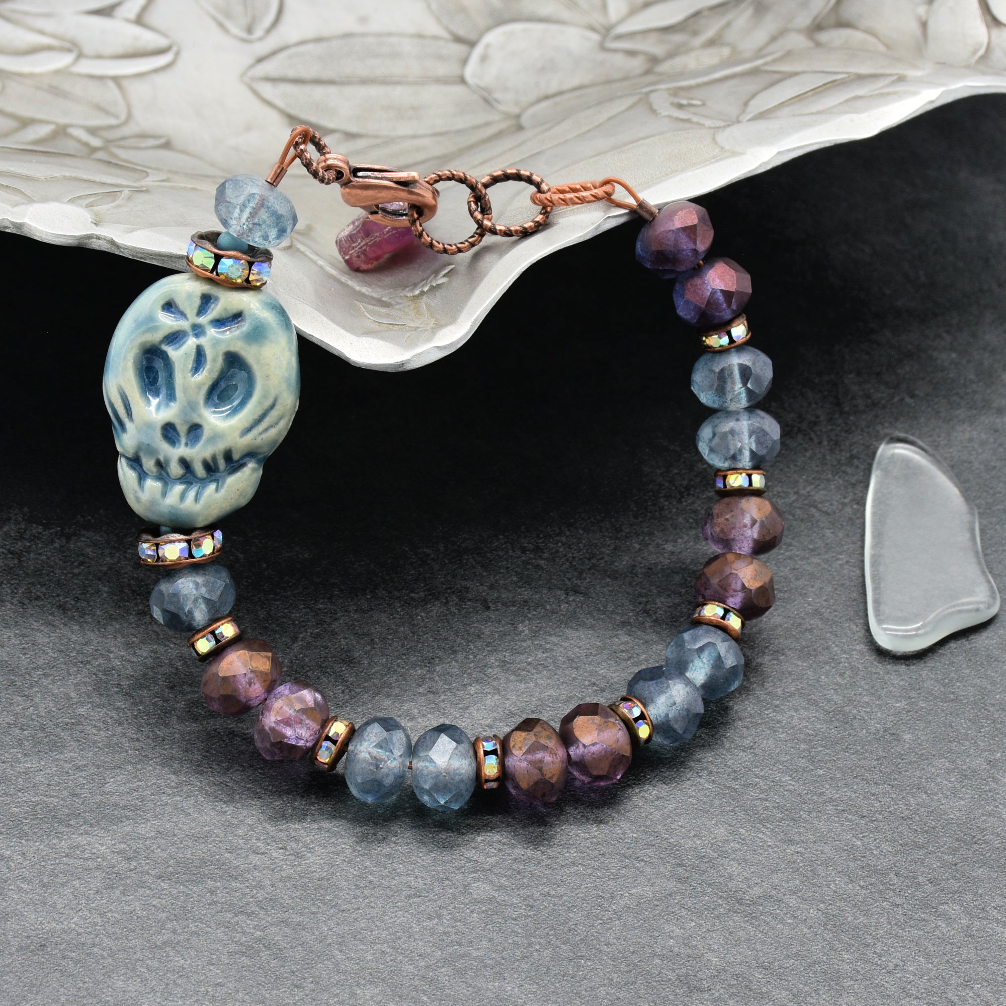 A beaded bracelet with a blue Dia de los Muertos sugar skull, faceted beads in blue & purple, and copper-toned metal accents, displayed on a dark gray surface with a silver tray - made with love in Colorado by Ruby Acorn.