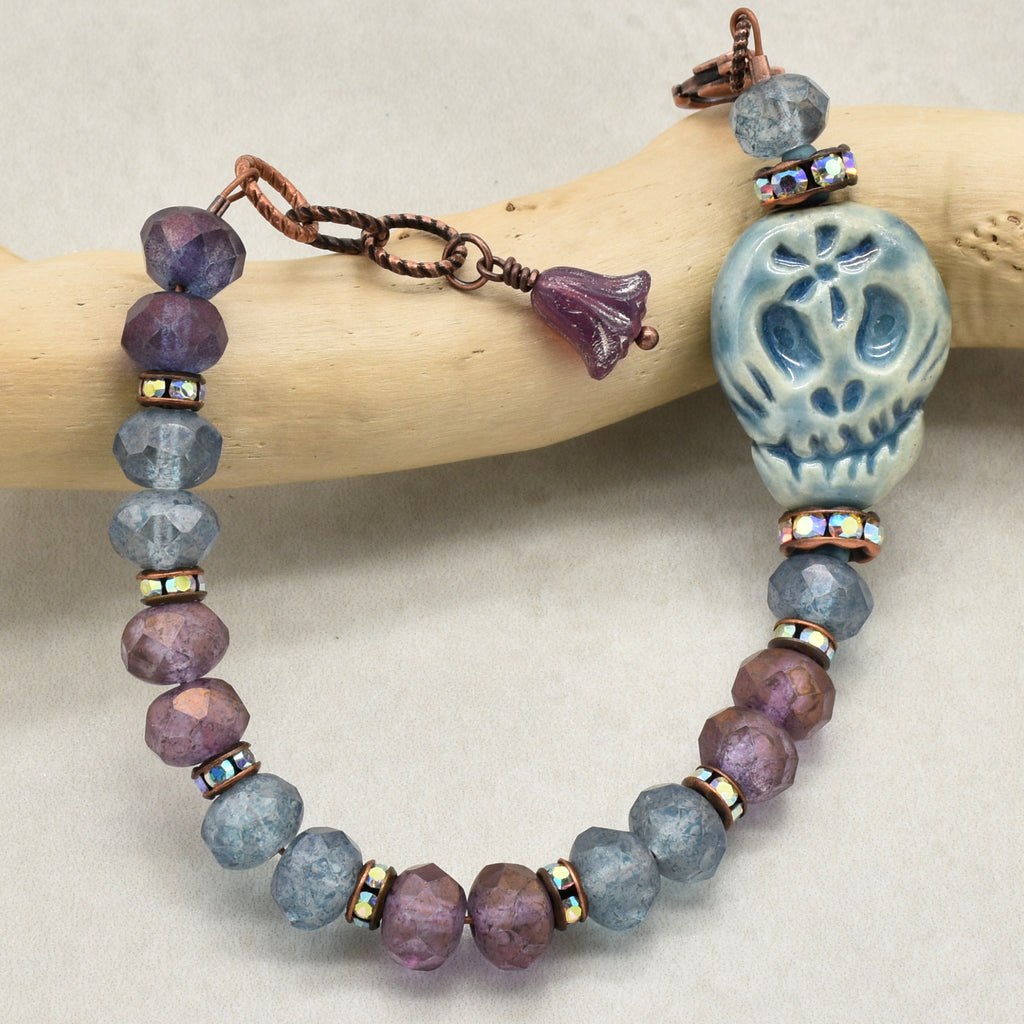 A beaded bracelet with faceted purple and blue-gray beads, a blue Day of the Dead sugar skull bead, iridescent spacer beads, and a copper-toned clasp, displayed on a curved wooden branch against a neutral beige background - made with love in Colorado by Ruby Acorn.