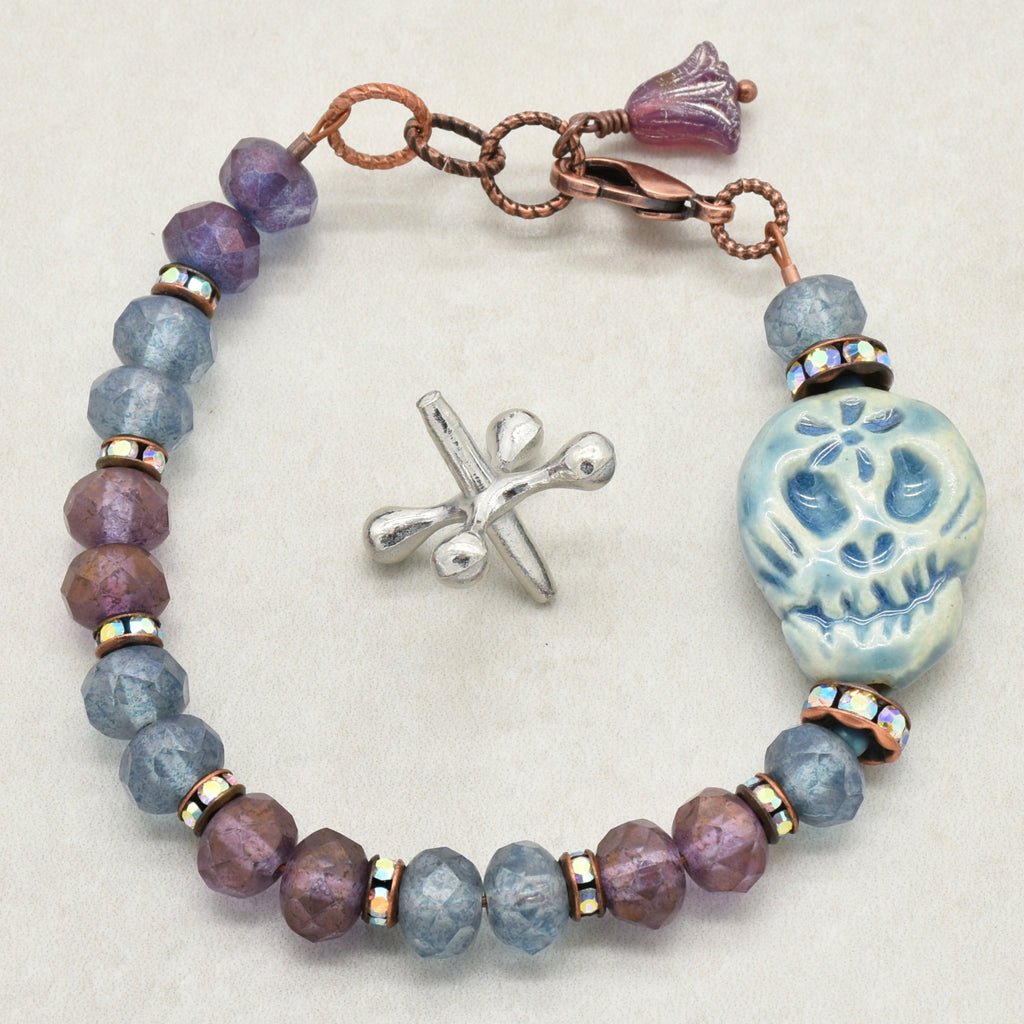 A beaded bracelet with faceted purple and slate blue beads, a blue Day of the Dead sugar skull charm, and a copper-toned lobster claw, on a light beige background - made with love in Colorado by Ruby Acorn.