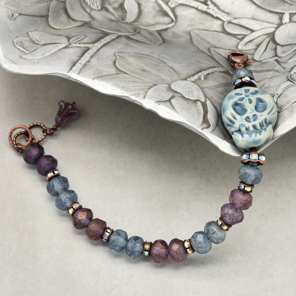 A beaded bracelet with faceted purple and slate-blue beads, accented by small iridescent spacers and a blue Dia de los Muertos sugar skull, displayed on a textured silver tray - made with love in Colorado by Ruby Acorn.