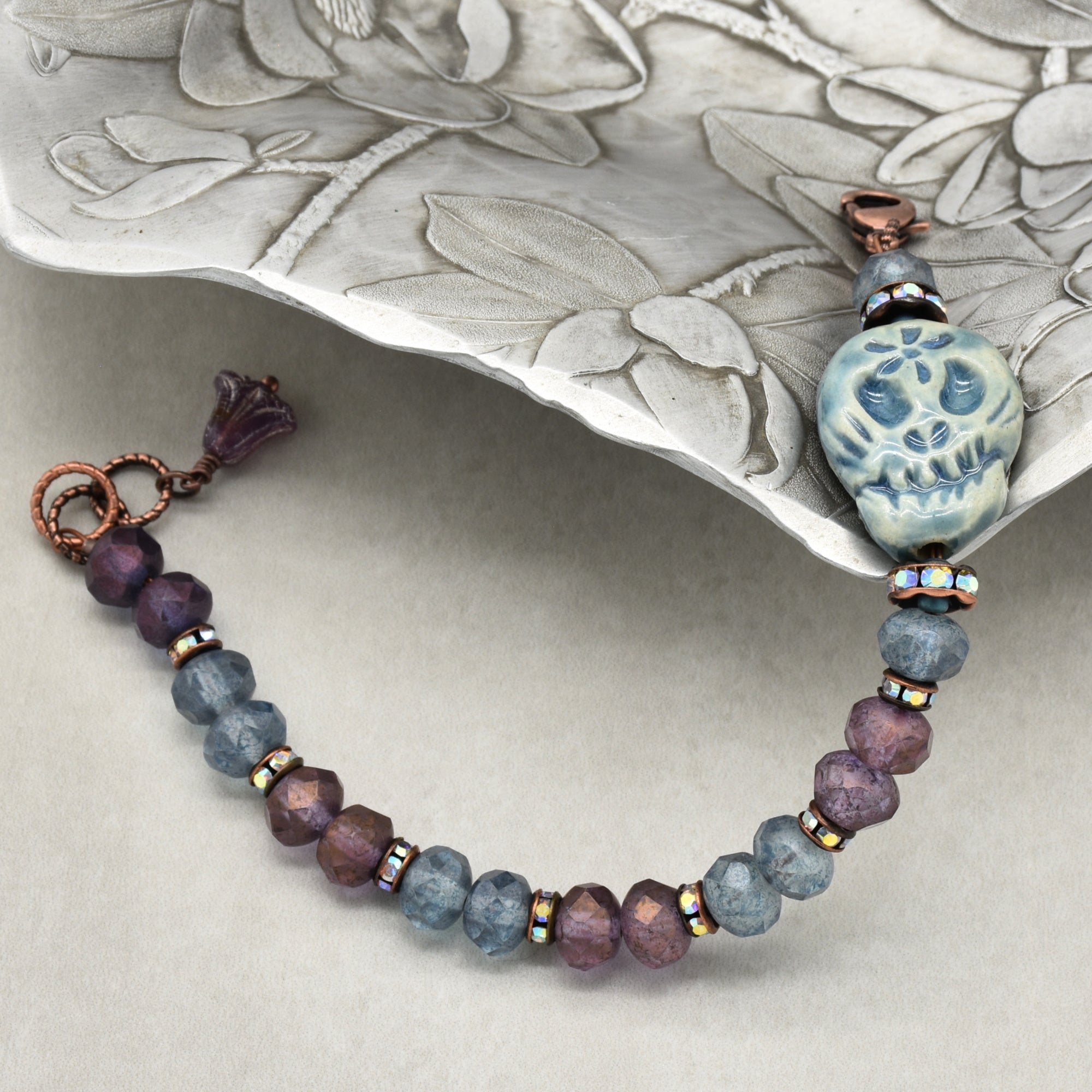 A beaded bracelet with faceted purple and slate-blue beads, accented by small iridescent spacers and a blue Dia de los Muertos sugar skull, displayed on a textured silver tray - made with love in Colorado by Ruby Acorn.