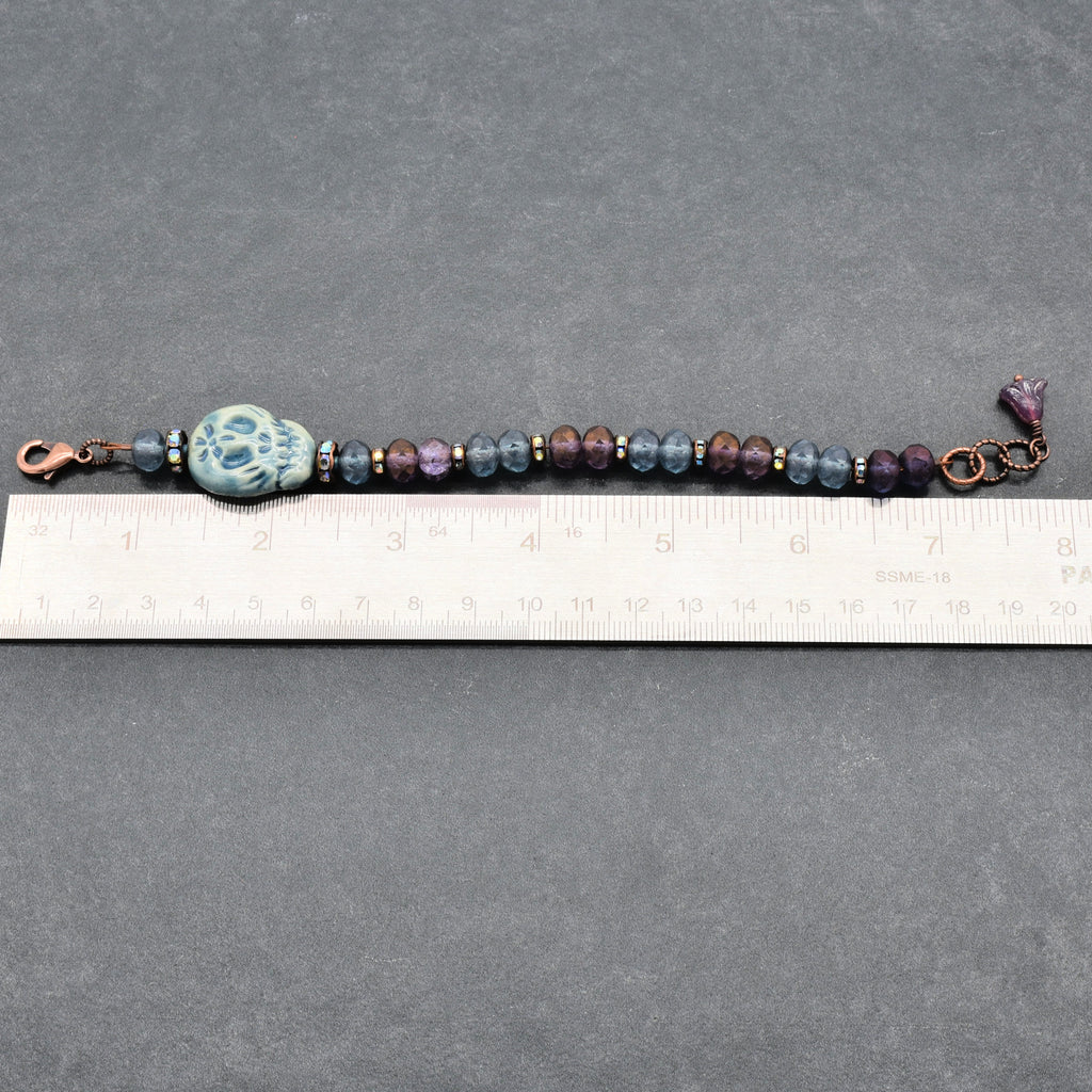 A beaded bracelet with a blue Day of the Dead sugar skull, dark purple and blue beads, and copper-toned metal accents, shown on a gray background with a ruler for scale - made with love in Colorado by  Ruby Acorn.