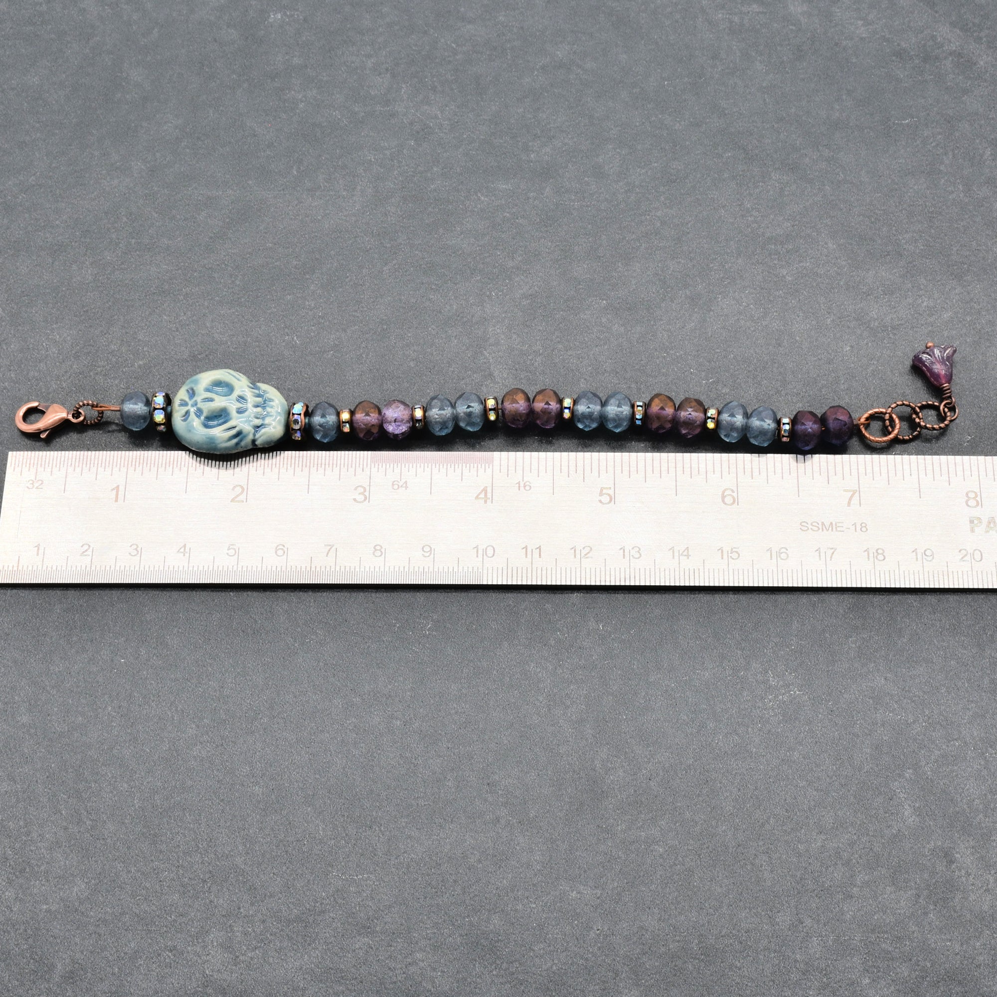 A beaded bracelet with a blue Day of the Dead sugar skull, dark purple and blue beads, and copper-toned metal accents, shown on a gray background with a ruler for scale - made with love in Colorado by  Ruby Acorn.