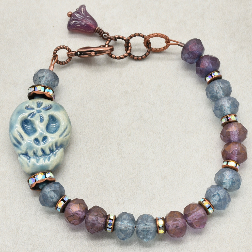 A beaded bracelet featuring a Day of the Dead sugar skull bead, faceted beads in lavender-purple and dusty blue, and copper-toned metal spacers and clasp - made with love in Colorado by Ruby Acorn.