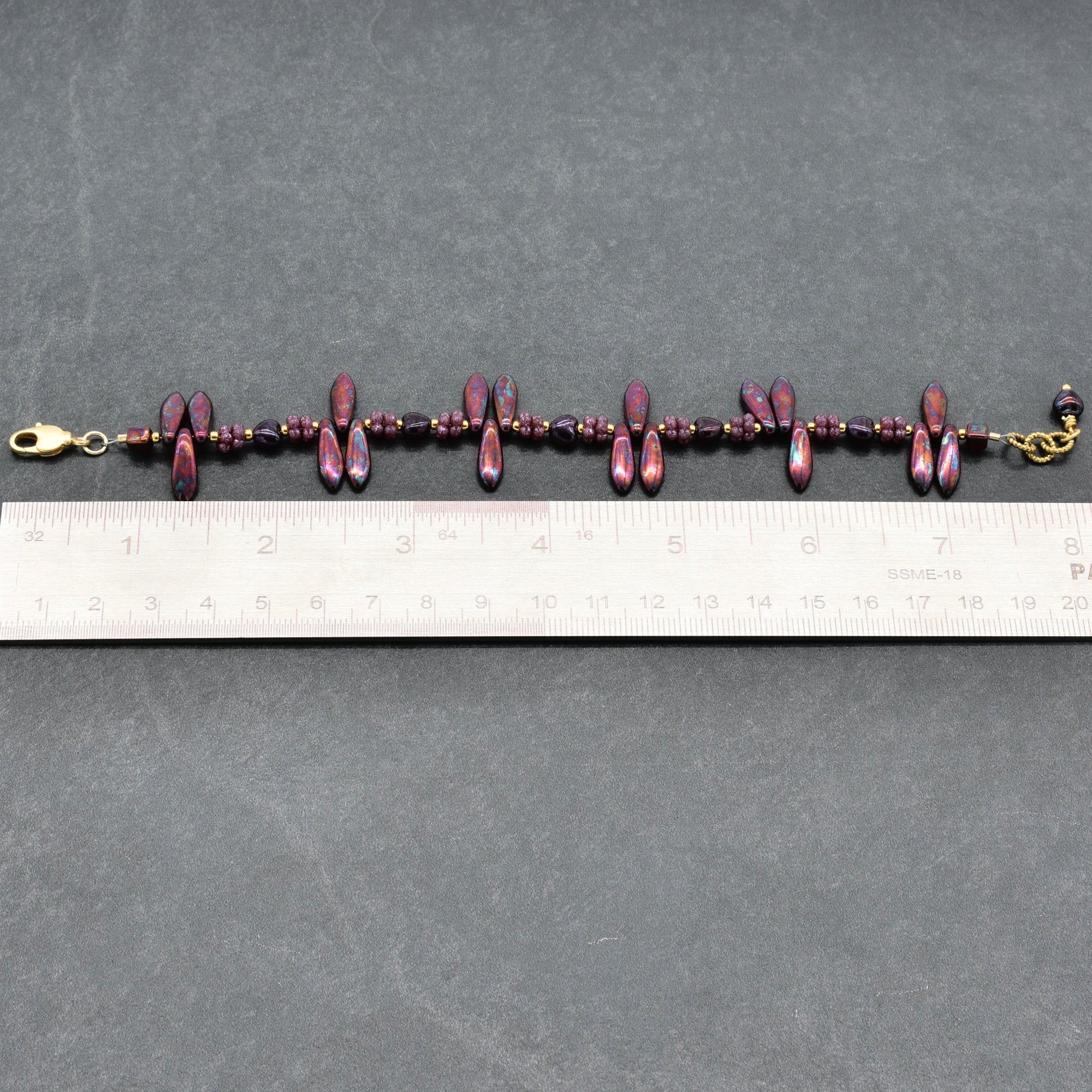 Bracelet with burgundy beads and gold clasp on a gray surface with a ruler for scale.
