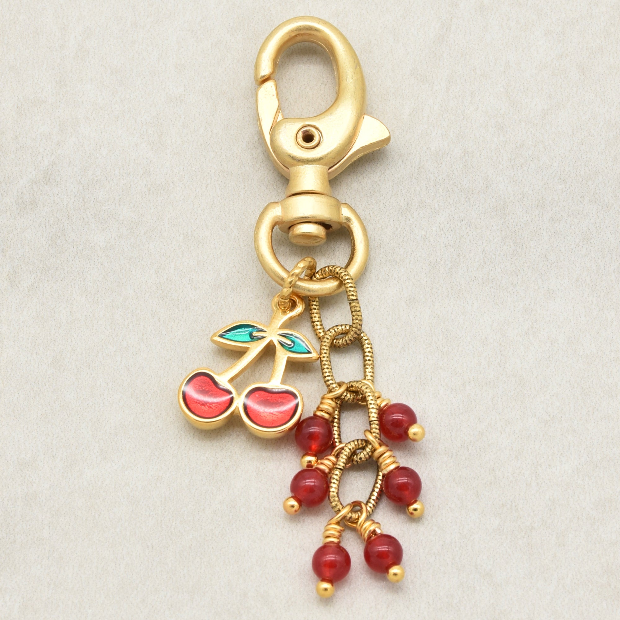 Gold-tone lobster clasp keychain charm featuring a cherry motif with red enamel and green leaves, alongside dangling red jade beads wrapped in gold wire, shown on a light textured background - made with love in Colorado by Ruby Acorn.