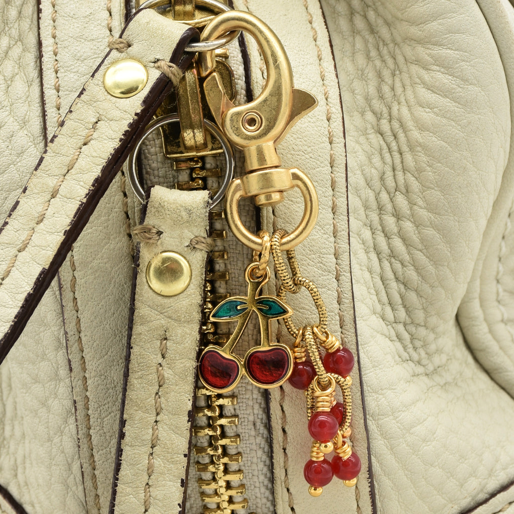 A gold-toned lobster clasp keychain or bag charm with dangling cherry and bead charms in red and green enamel, shown on a cream handbag - made with love in Colorado by Ruby Acorn.