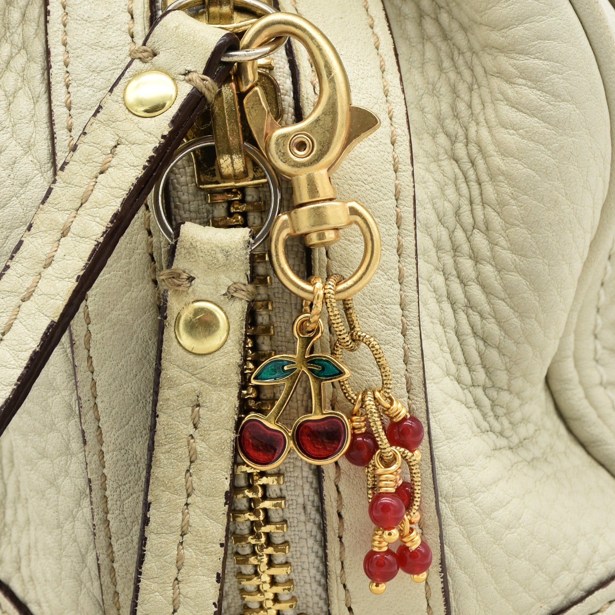 A gold-toned lobster clasp keychain or bag charm with dangling cherry and bead charms in red and green enamel, shown on a cream handbag - made with love in Colorado by Ruby Acorn.